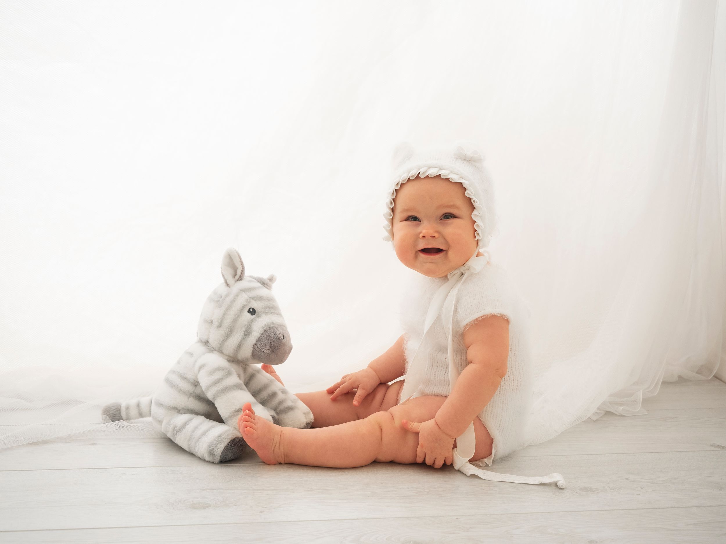baby girl in soft white vest and bonnet sits with soft toy zebra in front of white curtain