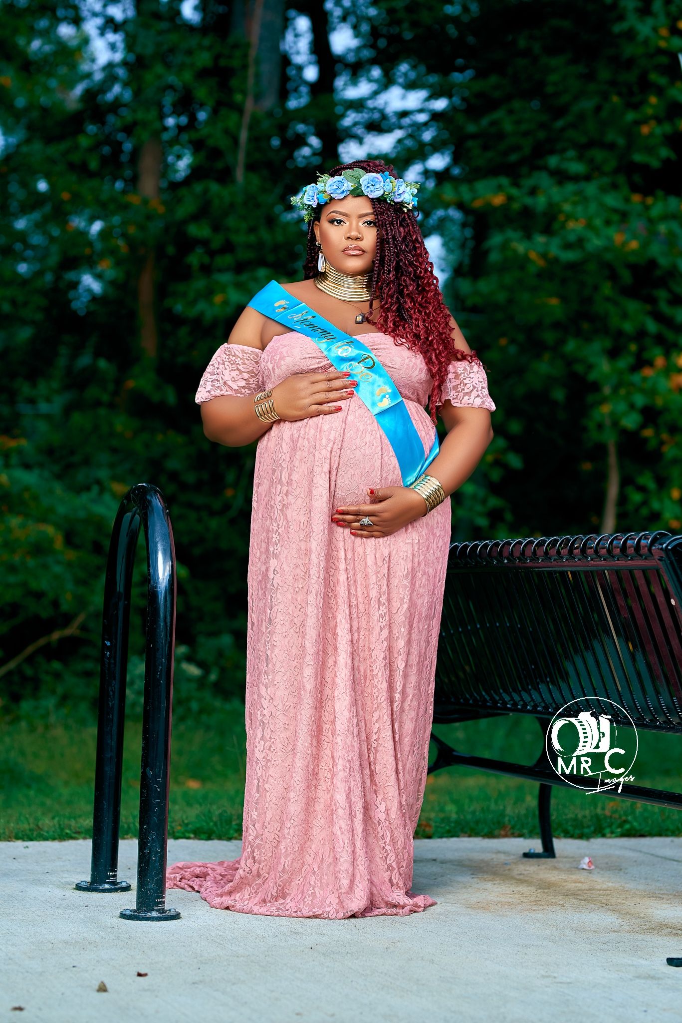 Maternity portrait in pink lace gown with blue sash against forest backdrop.