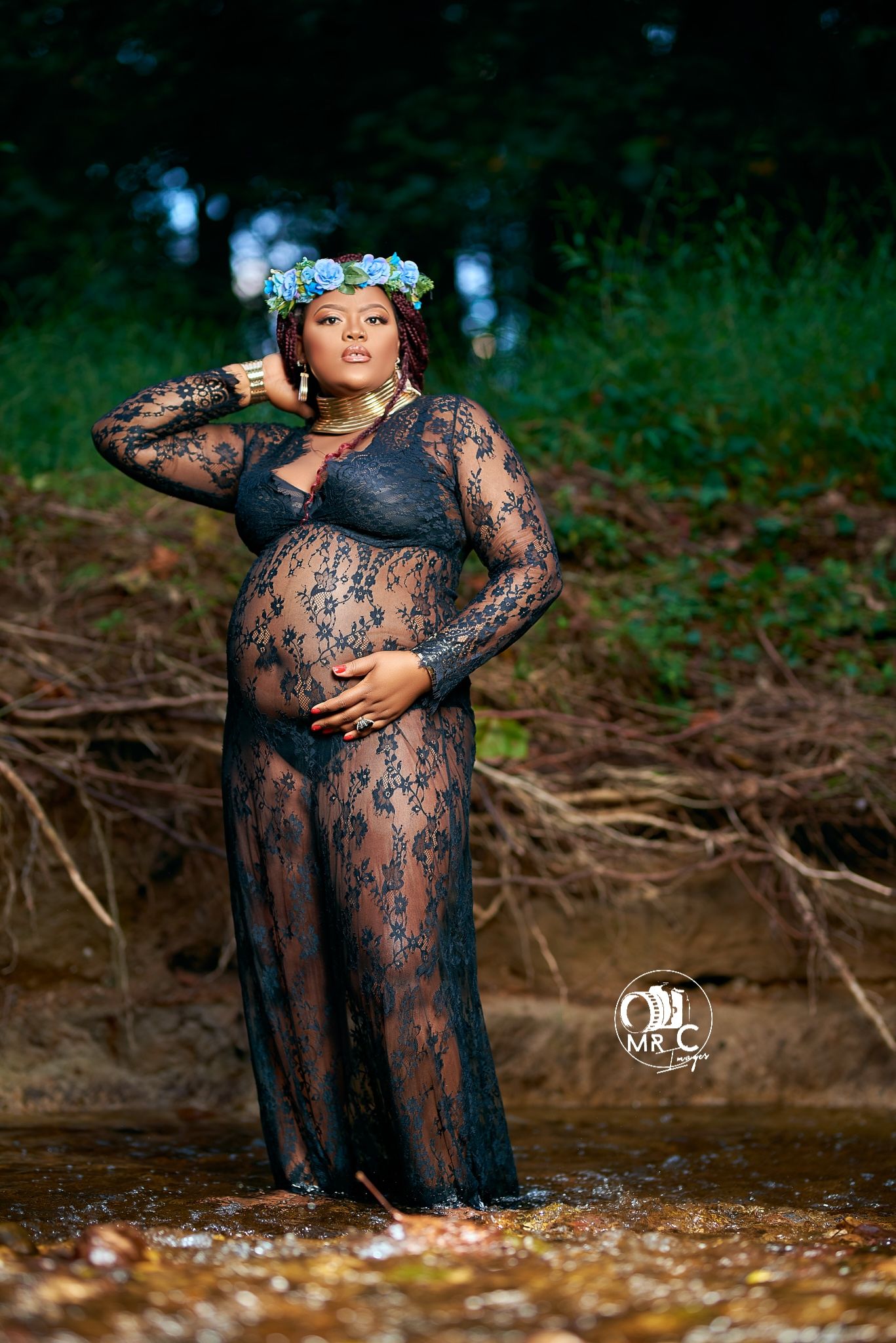 Artistic maternity photo in black lace dress by a scenic forest stream.