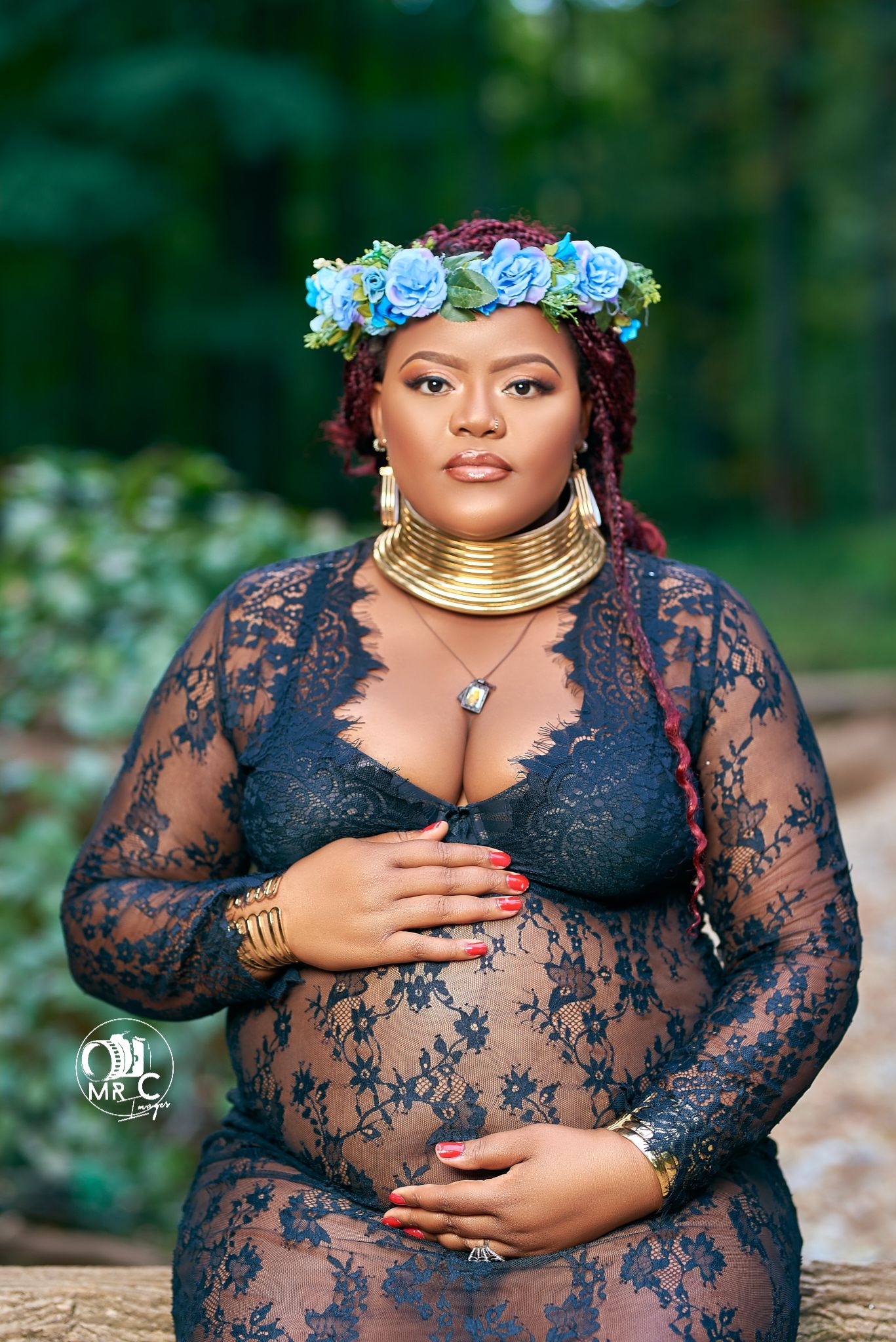 A maternity photo featuring a blue floral crown and gold statement necklace in an outdoor garden setting.
