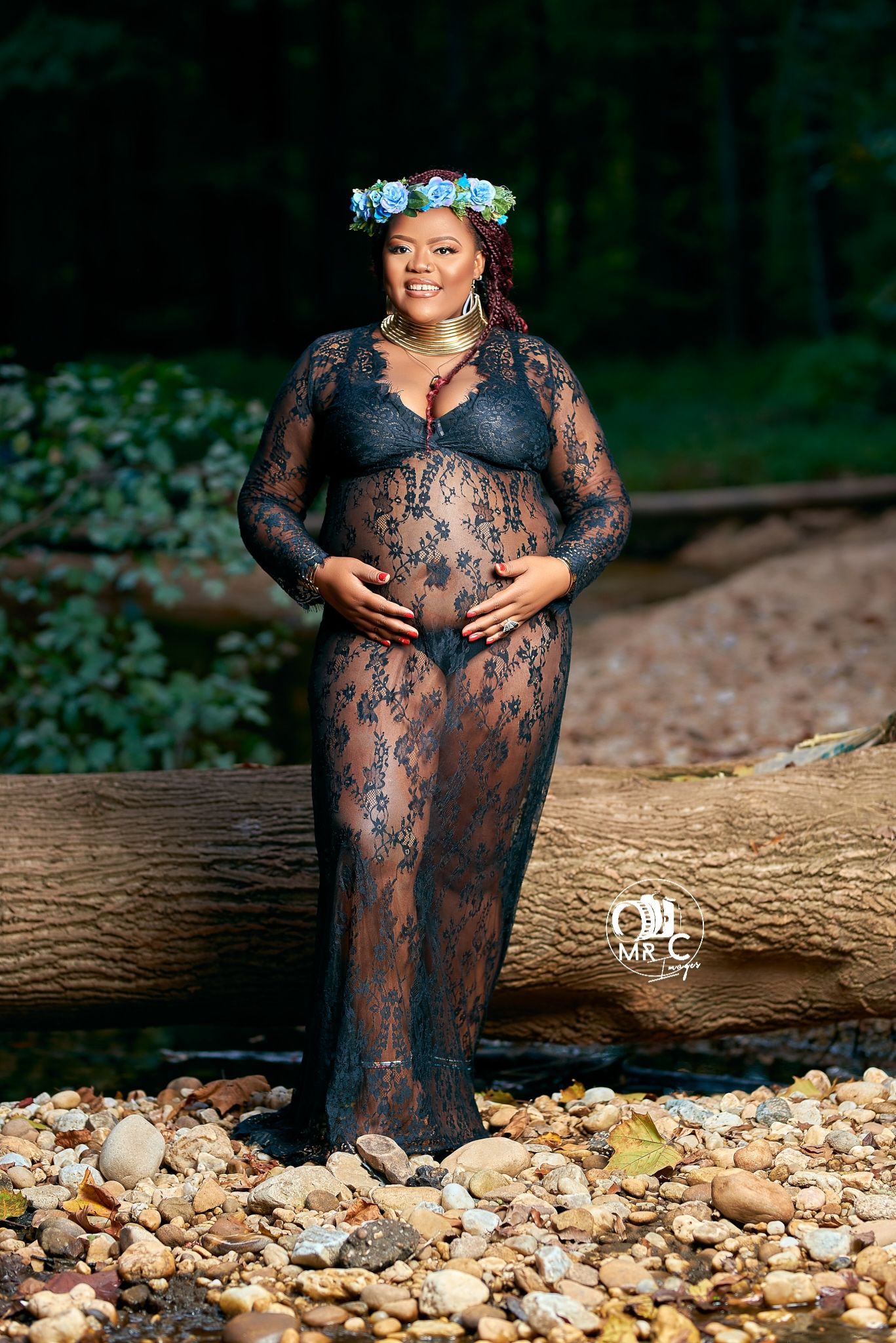 Artistic maternity portrait in black lace dress with floral crown standing on rocks by a stream.