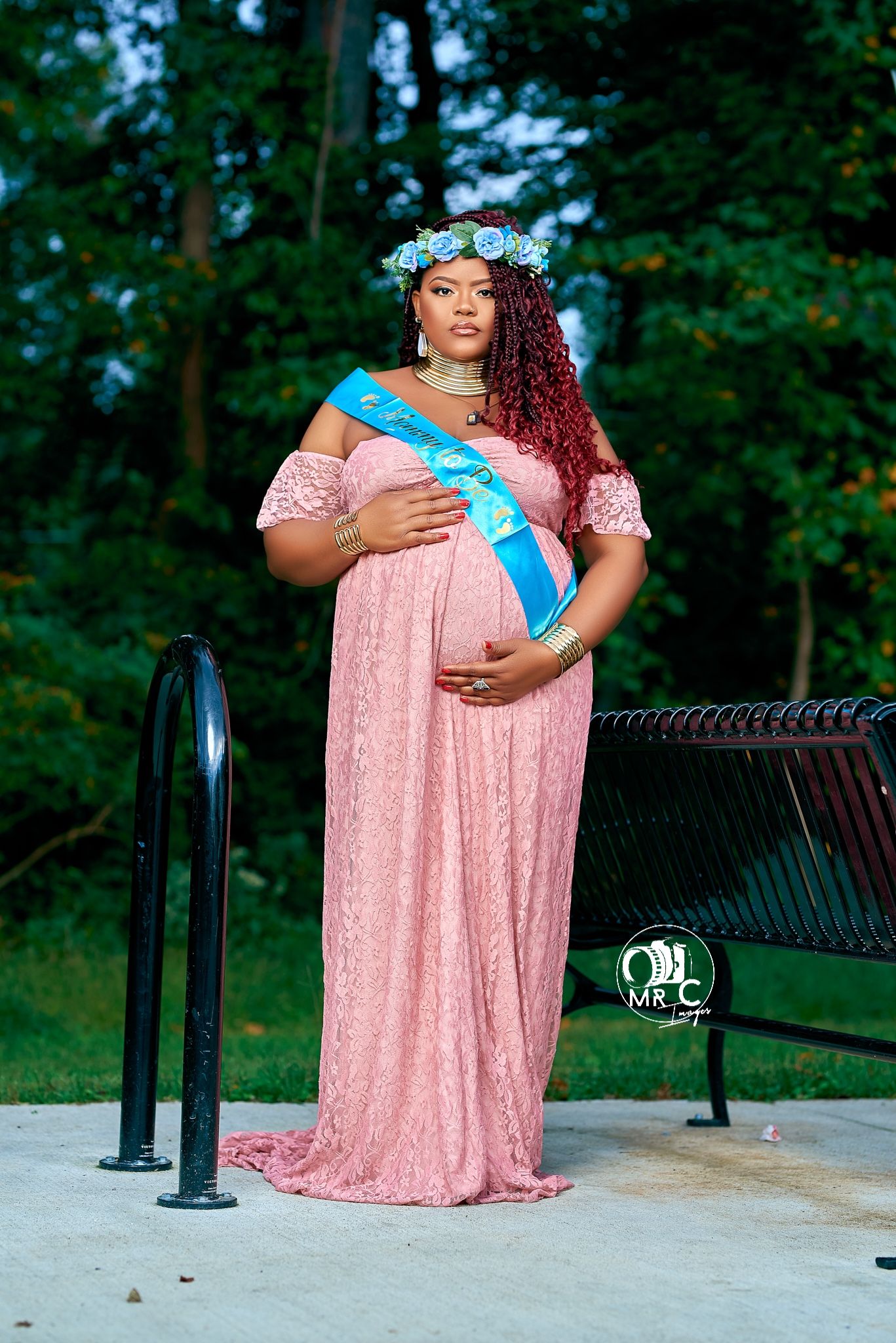 Expectant mother in pink lace gown and blue sash poses outdoors with floral crown.