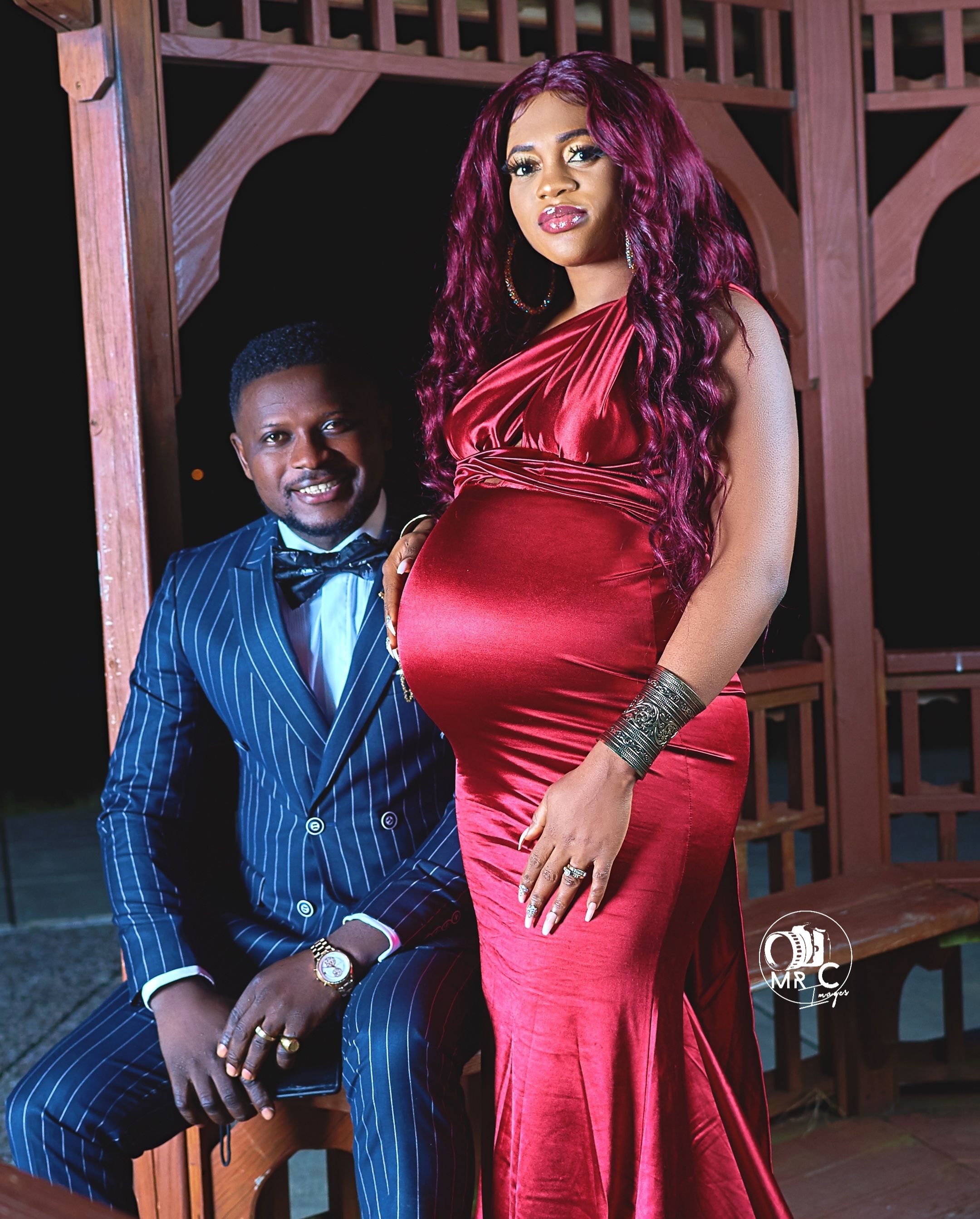 Glamorous maternity photoshoot with red satin gown and blue pinstripe suit in gazebo setting.