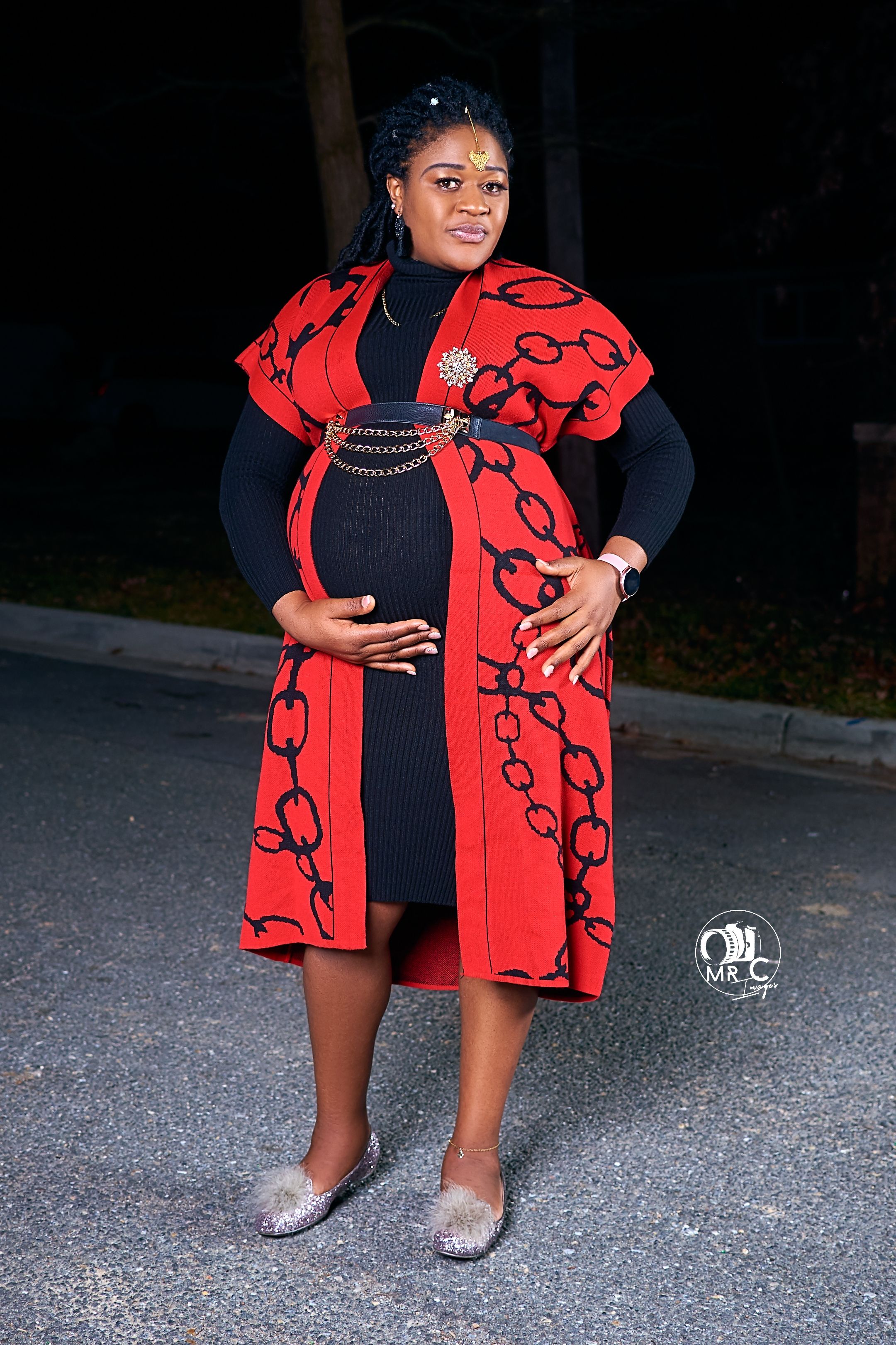 Stylish maternity portrait with a red chain-print coat dress and silver ballet flats on a dark street.