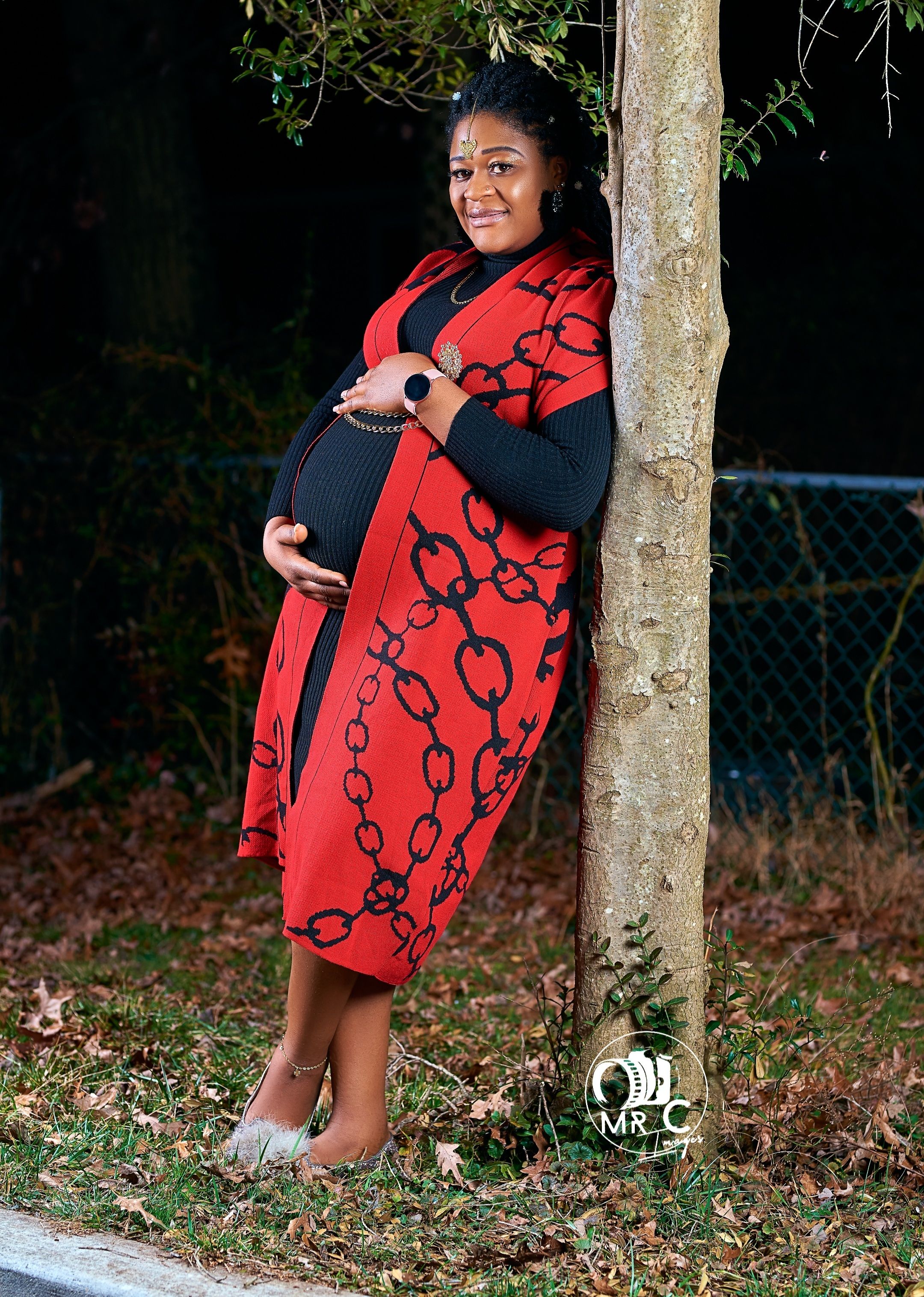 Maternity portrait showcasing a stylish red and black chain-print dress against an outdoor autumn backdrop.