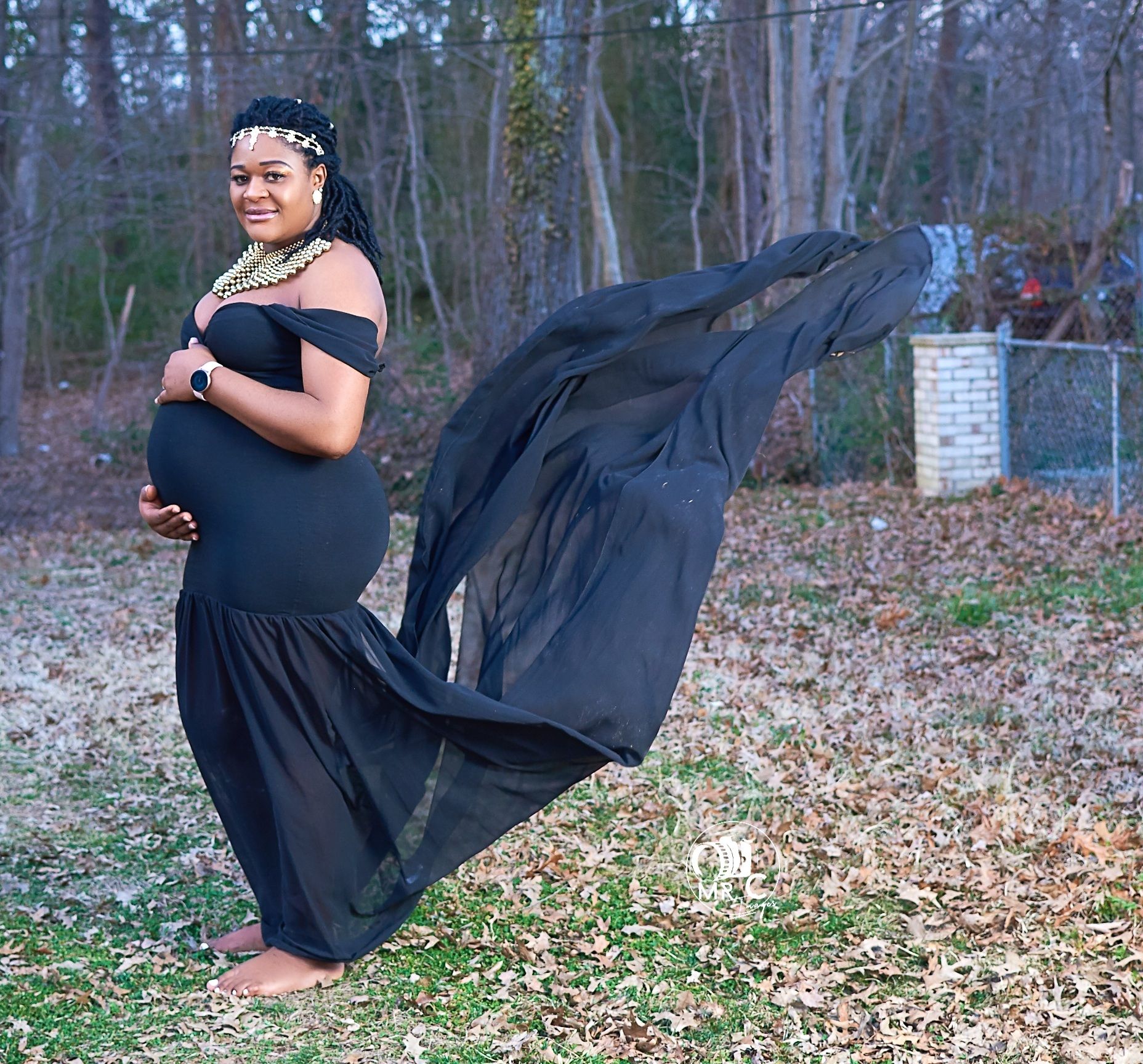 Elegant maternity photo featuring a flowing black dress with dramatic fabric movement in an outdoor wooded setting.