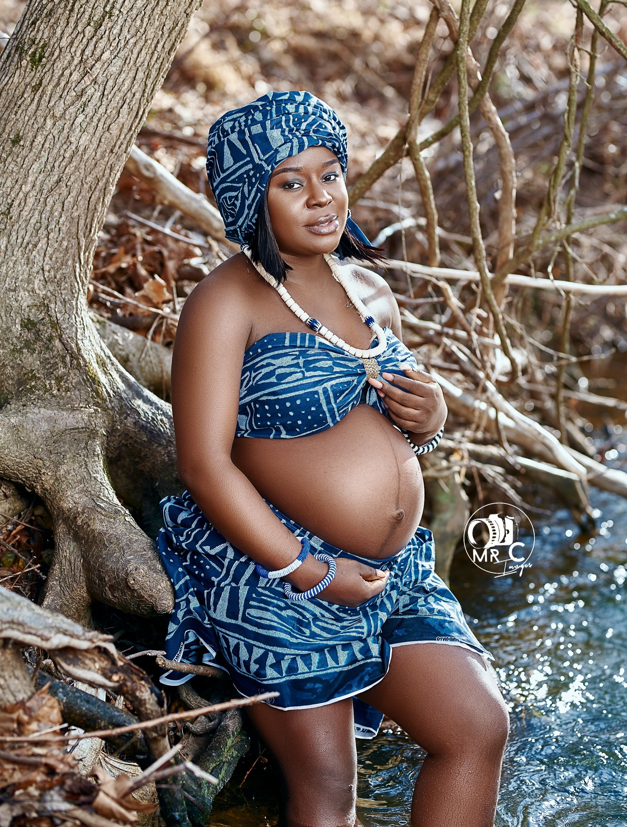Maternity portrait by tree wearing blue patterned two-piece outfit and matching headwrap.