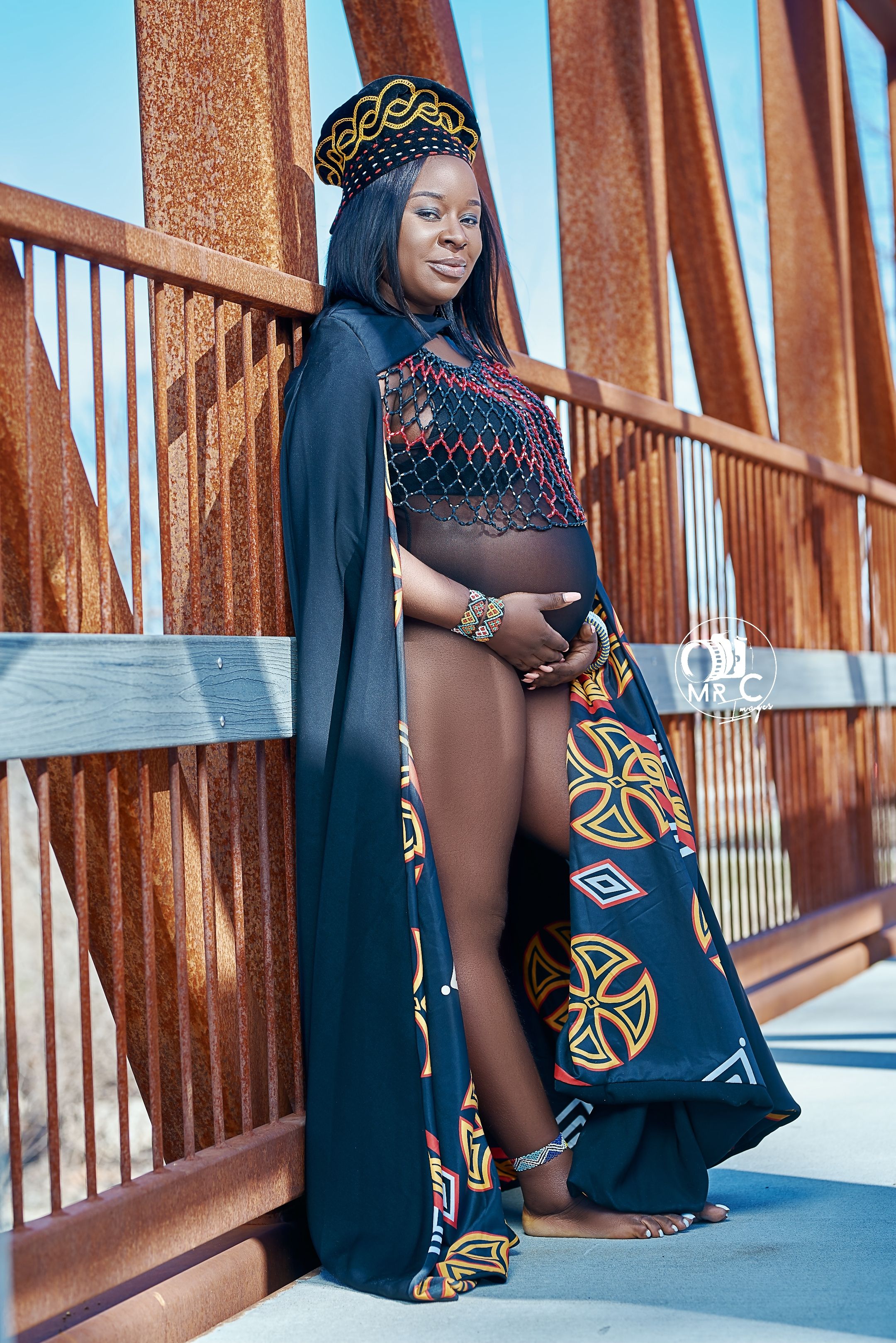 Dramatic maternity pose on bridge wearing black cape with geometric African print details.