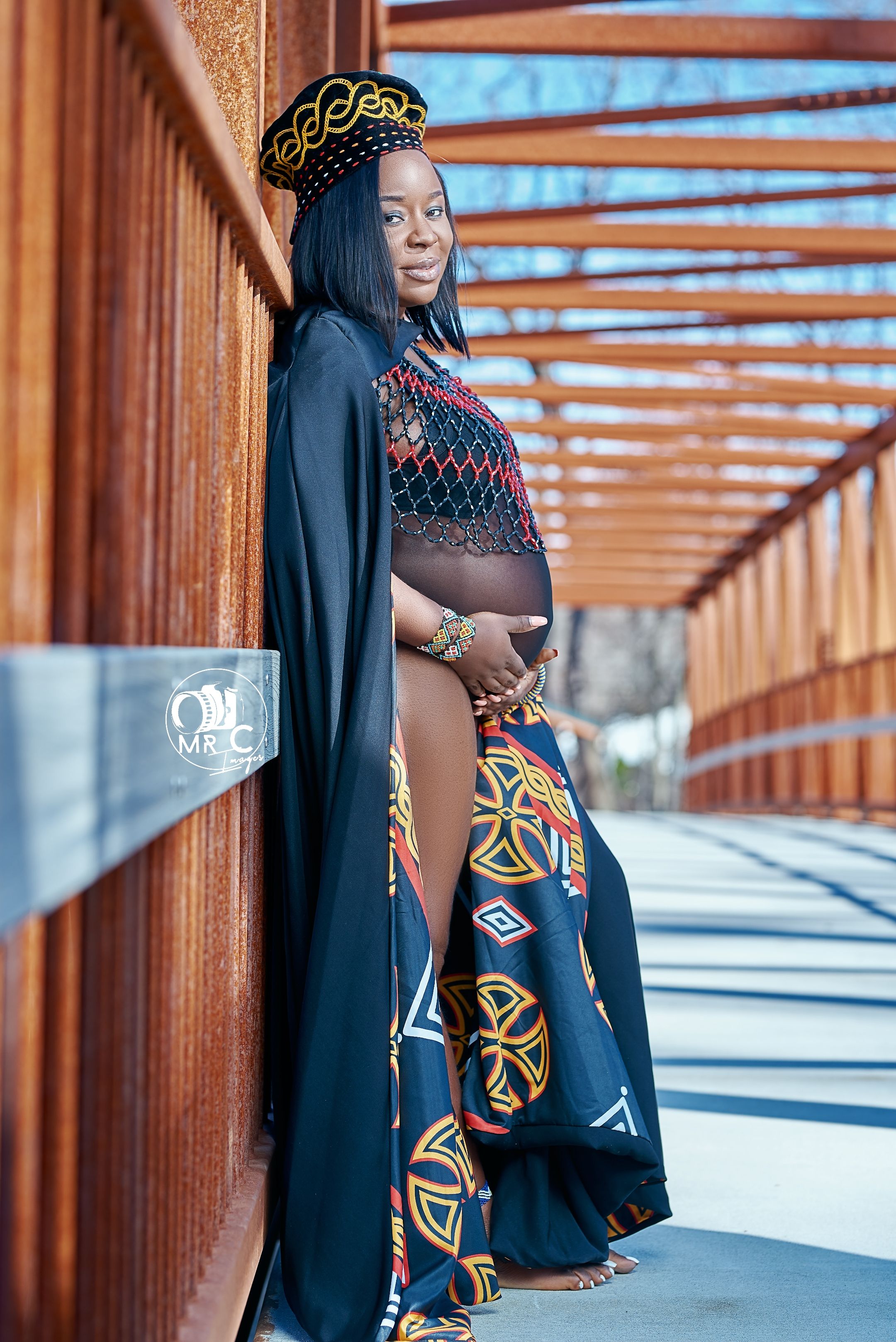 Artistic maternity portrait on rustic bridge with patterned cape and decorative headpiece.