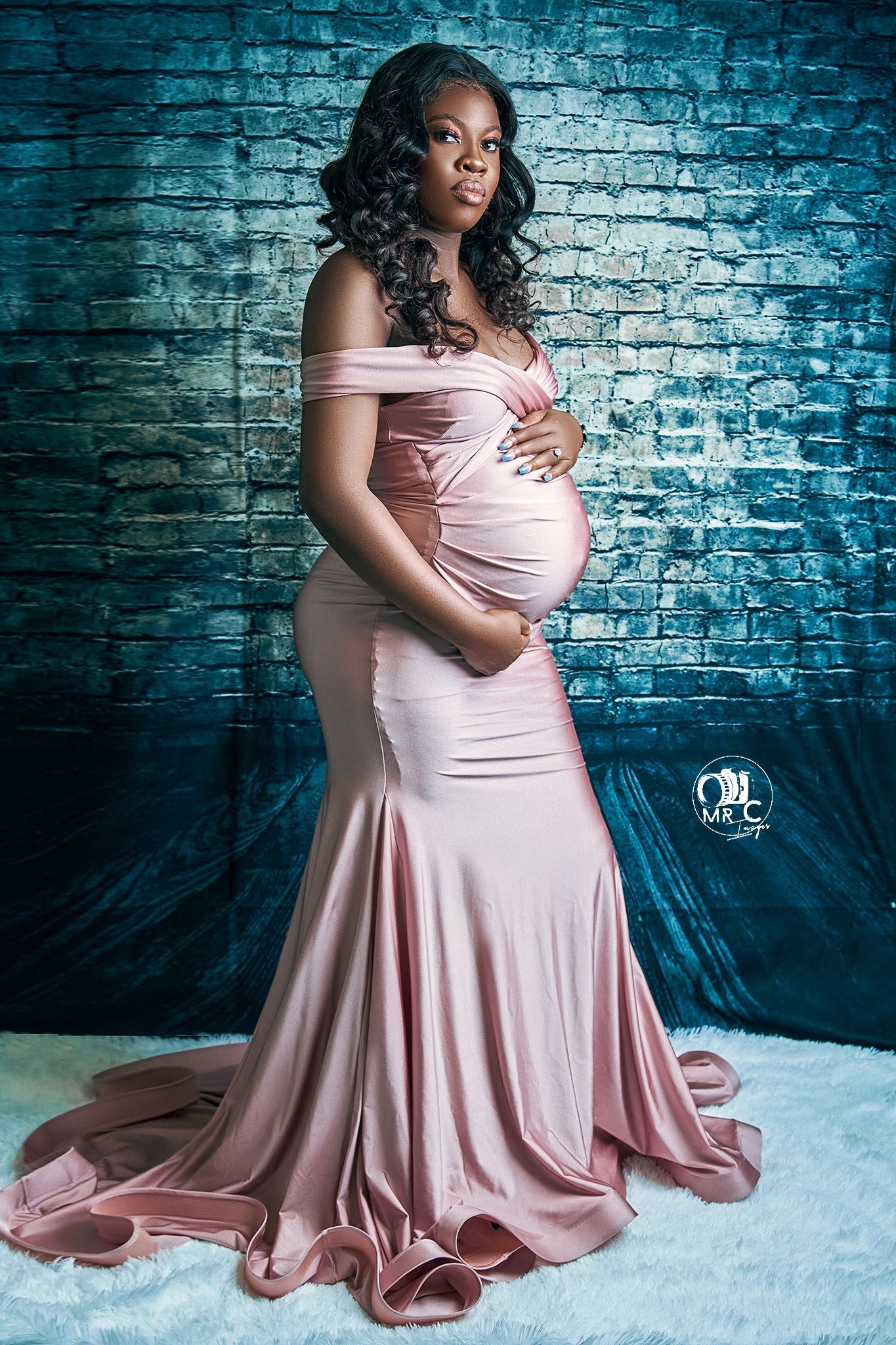 Elegant maternity portrait in pink off-shoulder gown against textured blue brick wall.