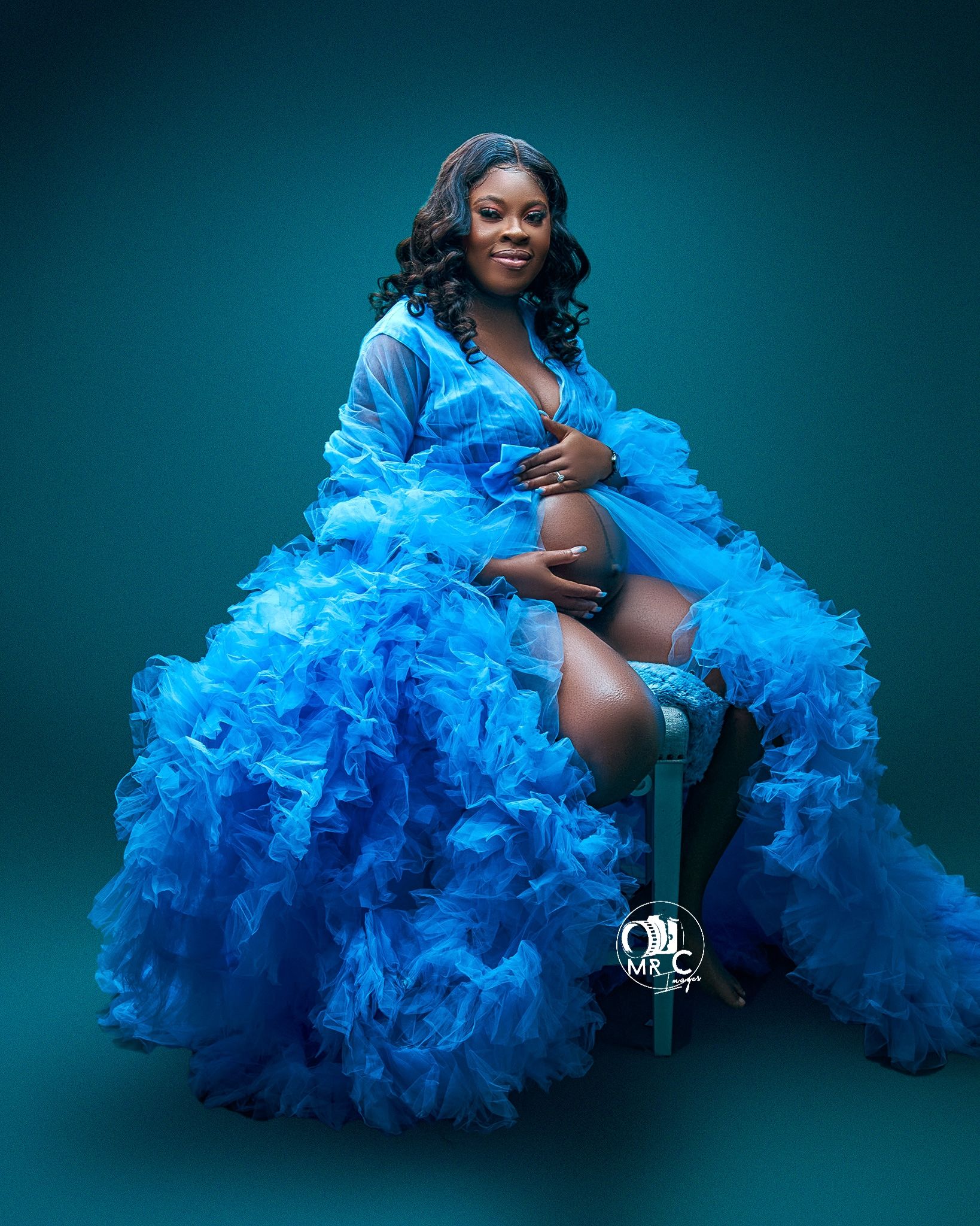 Glamorous maternity portrait featuring flowing blue ruffled gown against teal backdrop.