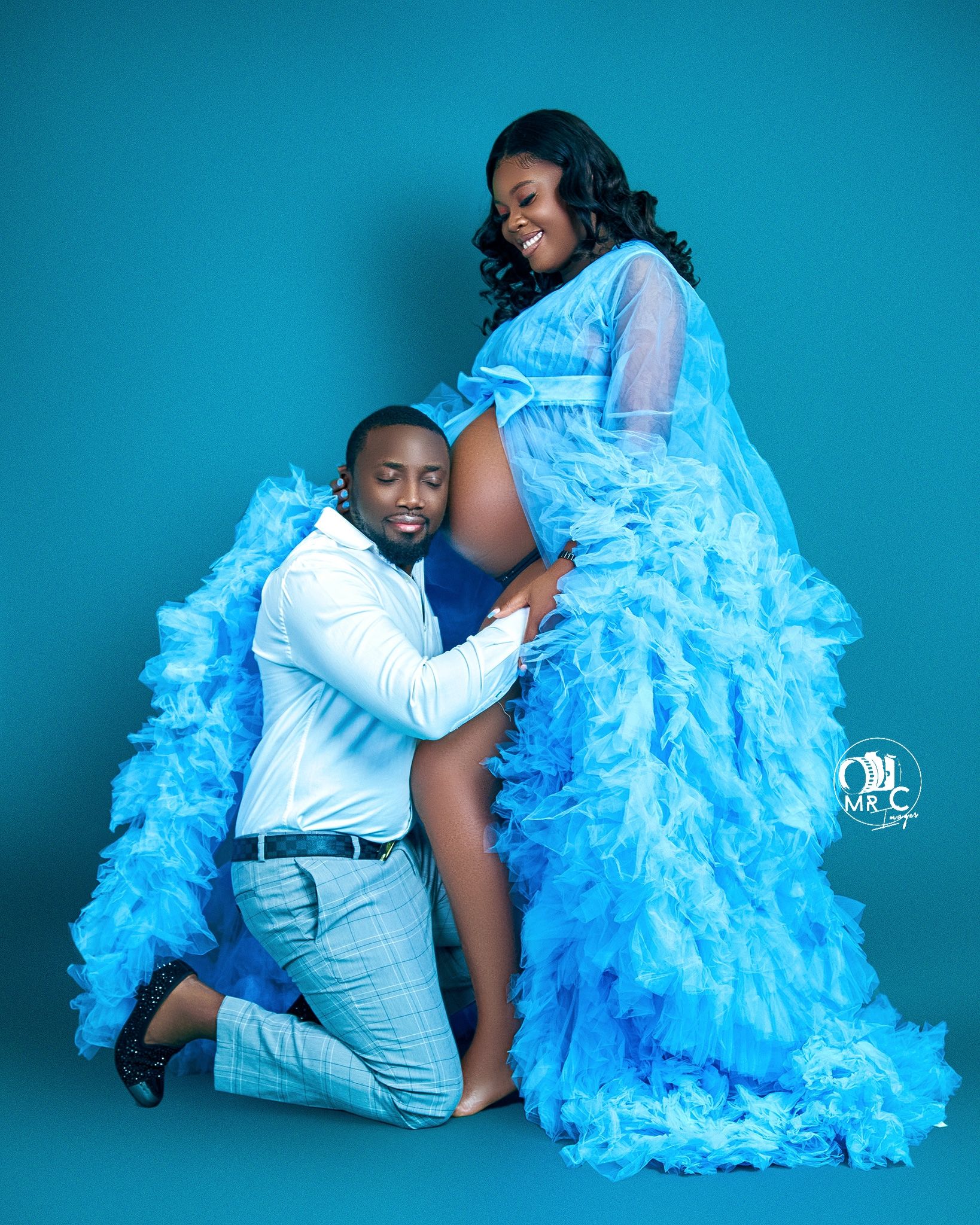 Intimate maternity portrait with couple in blue and white outfits against turquoise background.