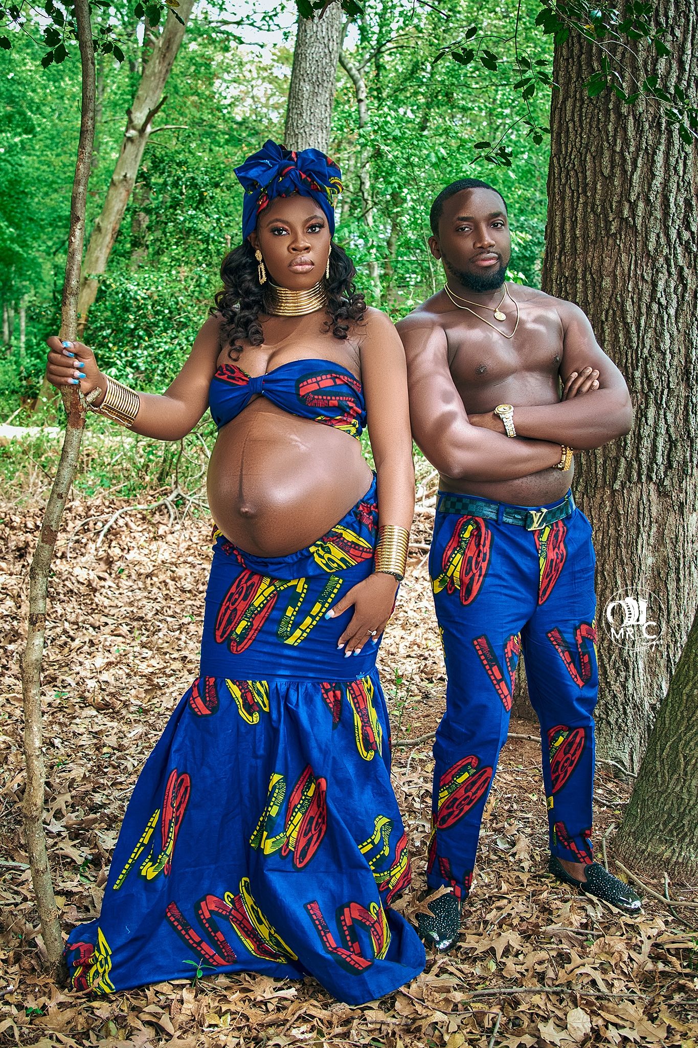 Couple in matching blue African print outfits pose in a forest setting during maternity photoshoot.