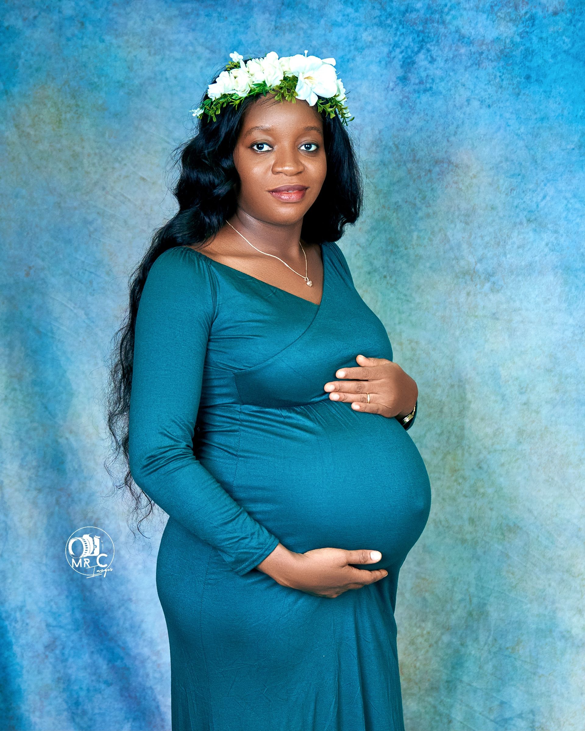Maternity portrait in teal dress with white floral crown against blue backdrop.
