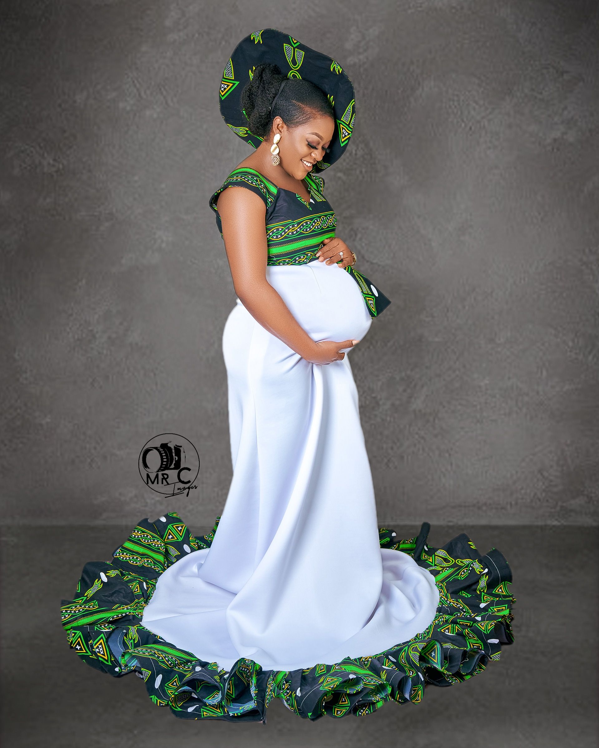 Elegant maternity portrait in white dress with green African print trim against grey background.