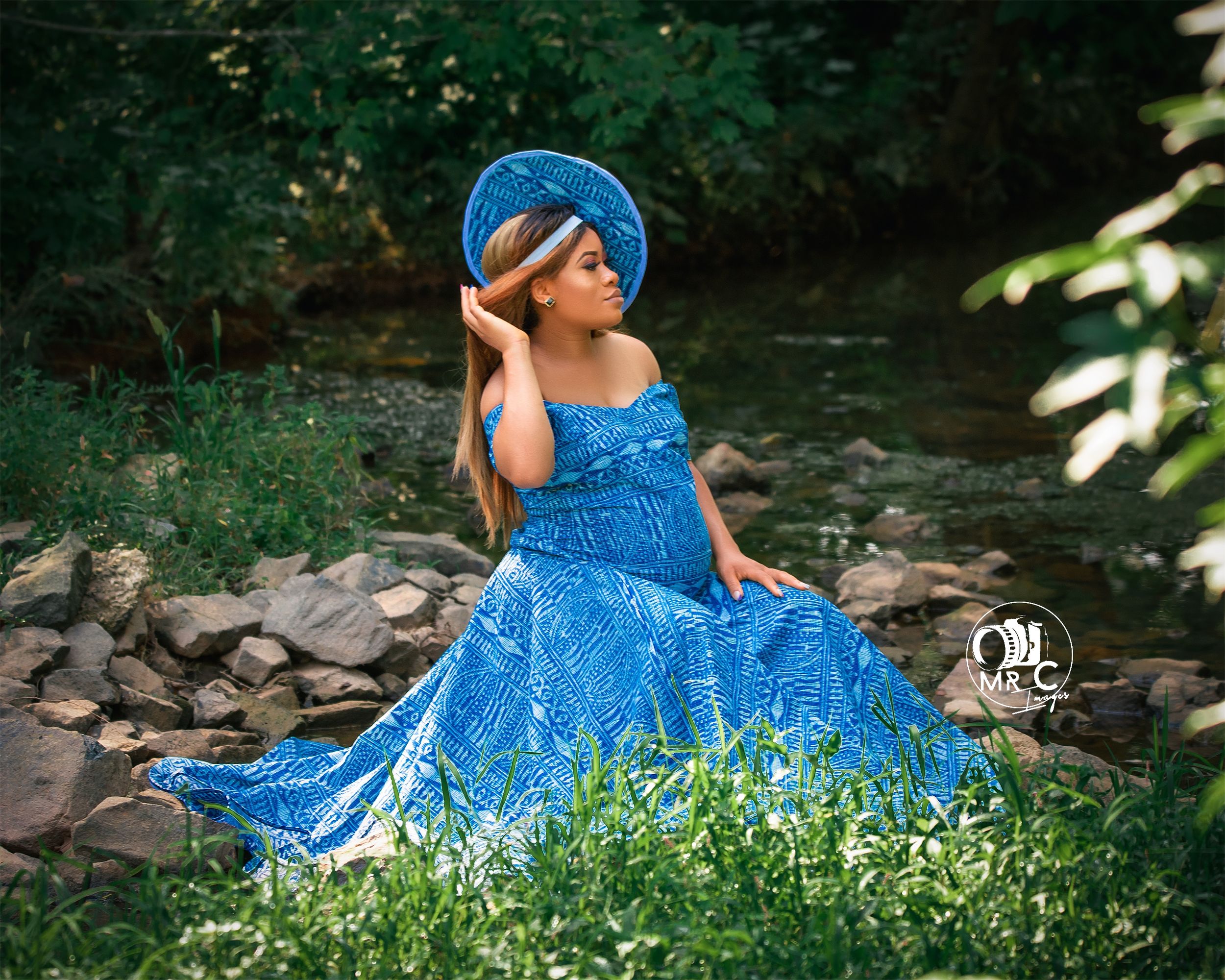 A flowing blue off-shoulder maternity gown styled with a matching hat in an outdoor setting.