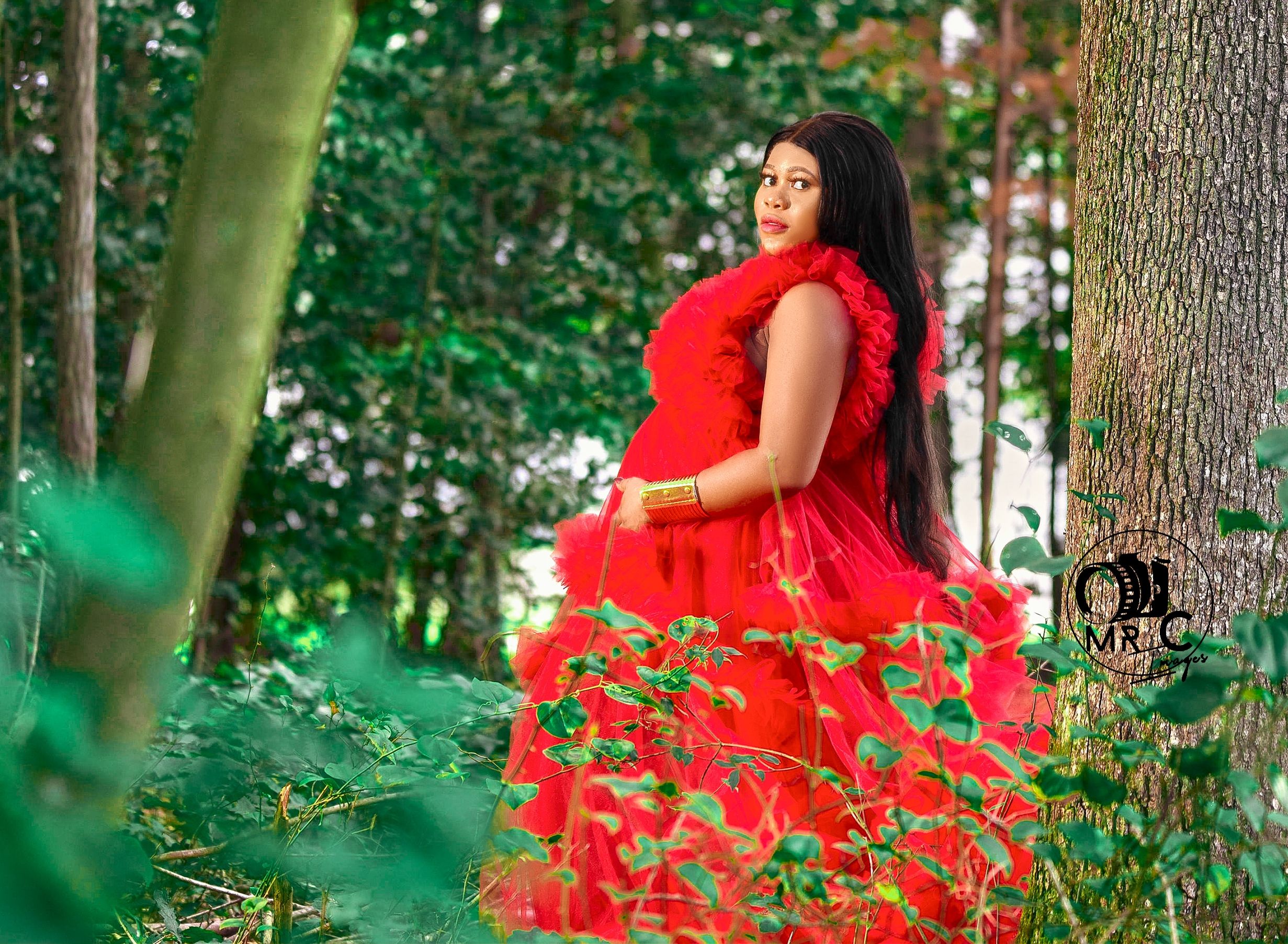 A dramatic red tulle gown photographed in a lush forest setting during a maternity shoot.