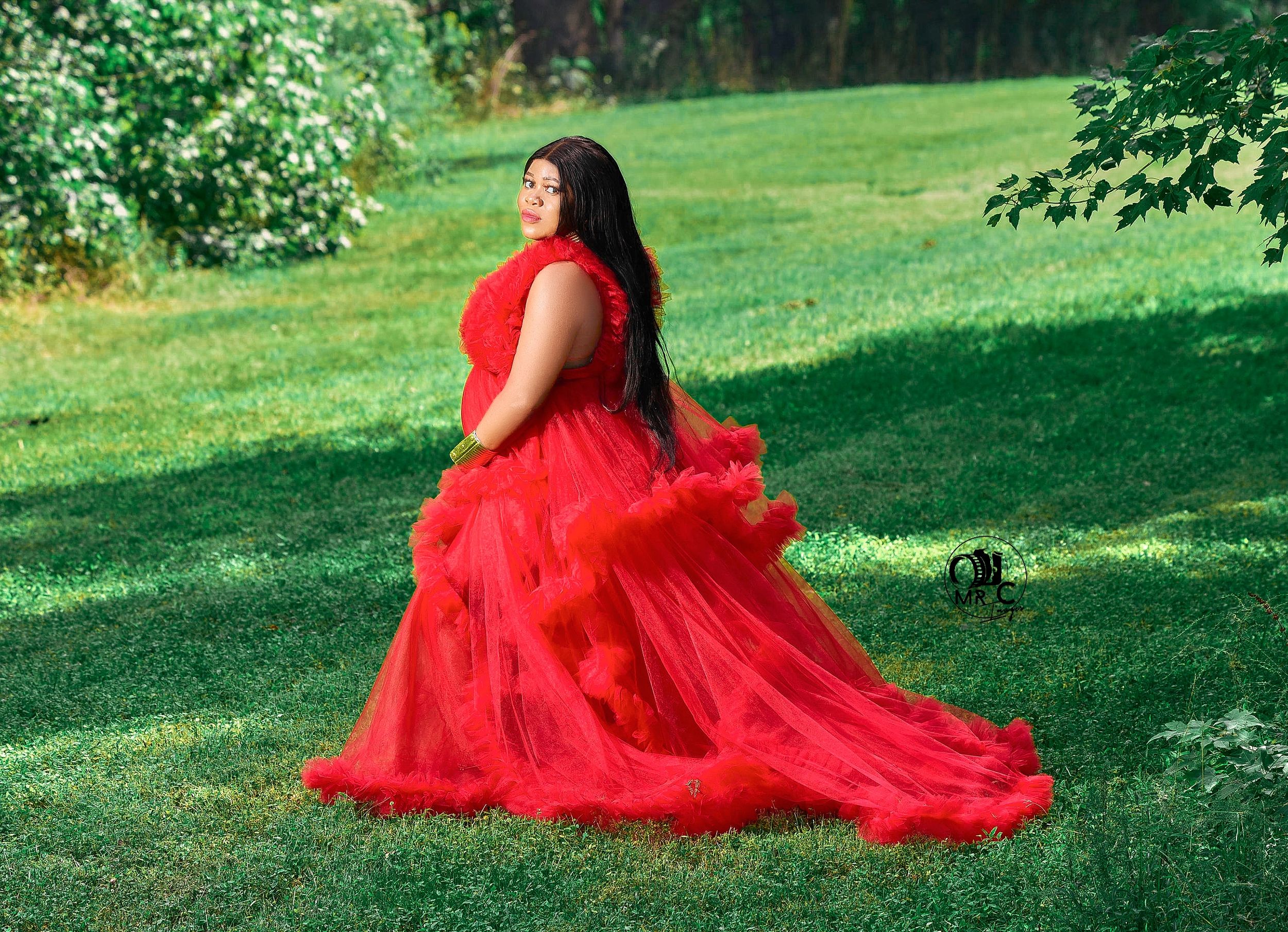 An elegant red tulle maternity gown photographed in a scenic outdoor garden setting.