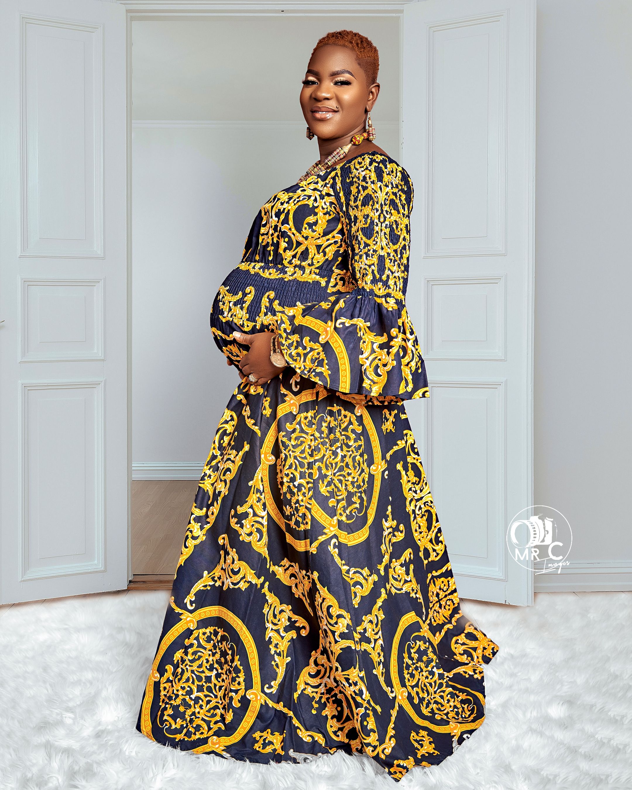 Maternity photo showcasing baroque-print navy and gold maxi dress against white interior.