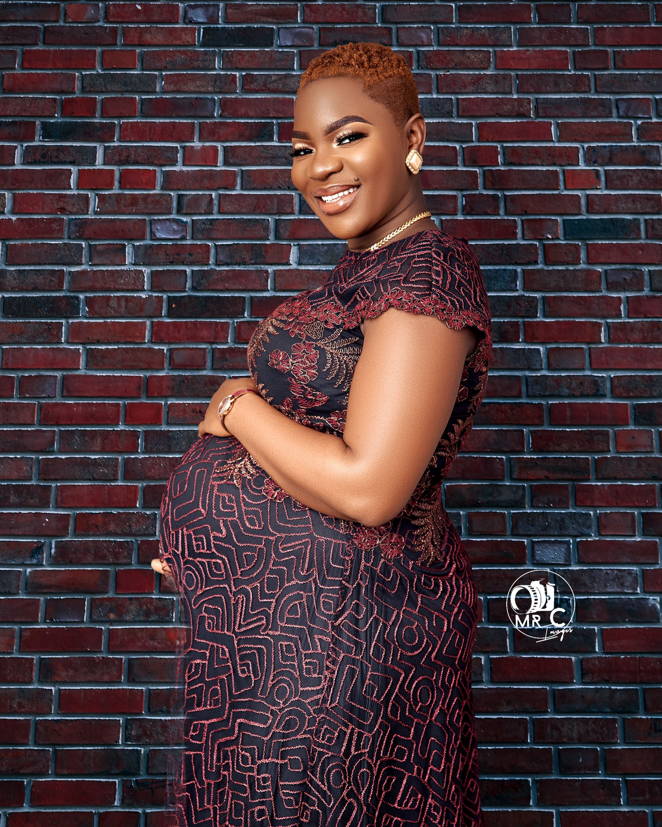 Maternity portrait against brick wall background wearing burgundy patterned dress.