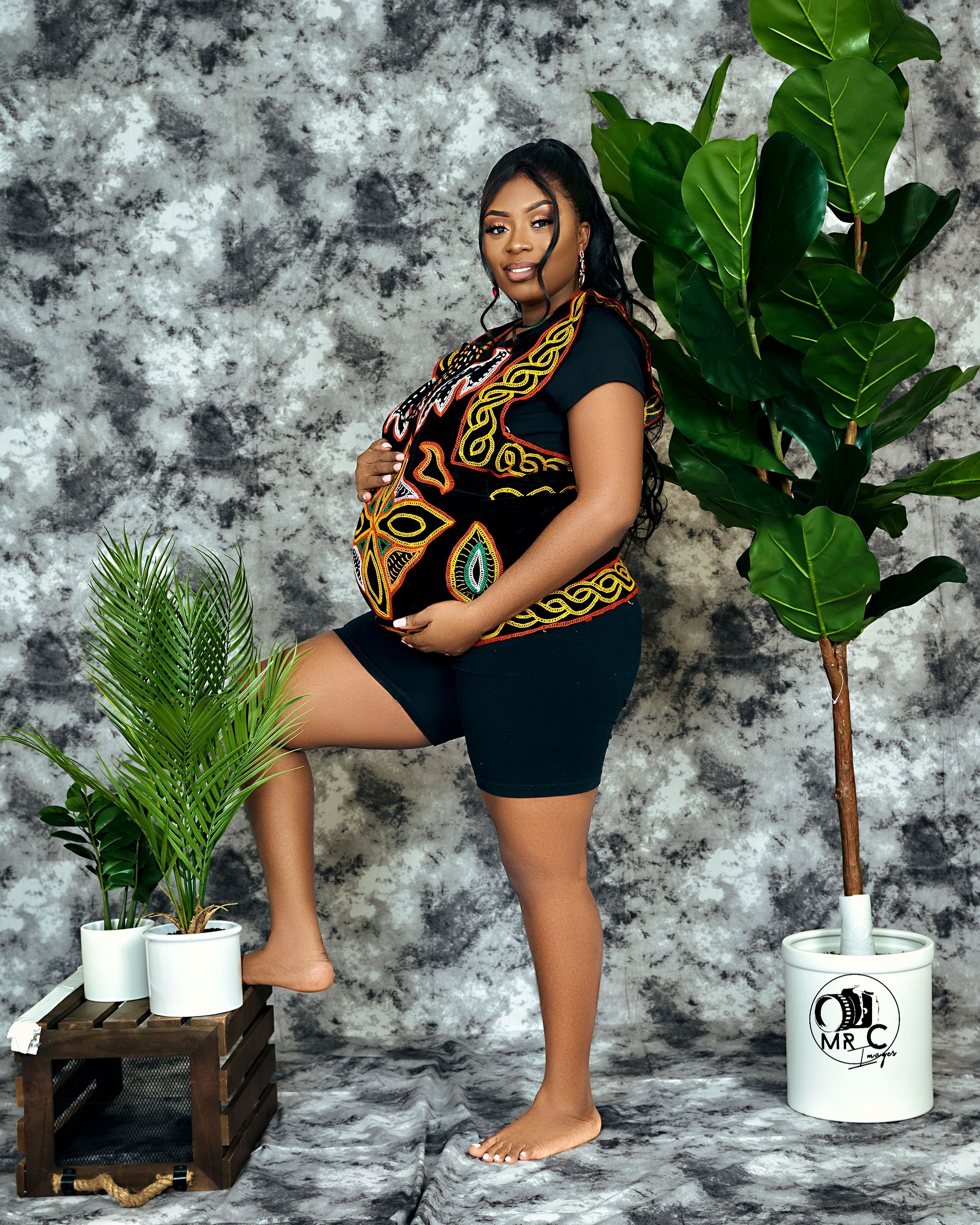 Maternity portrait in black and gold patterned outfit with tropical plants and wooden decor elements.