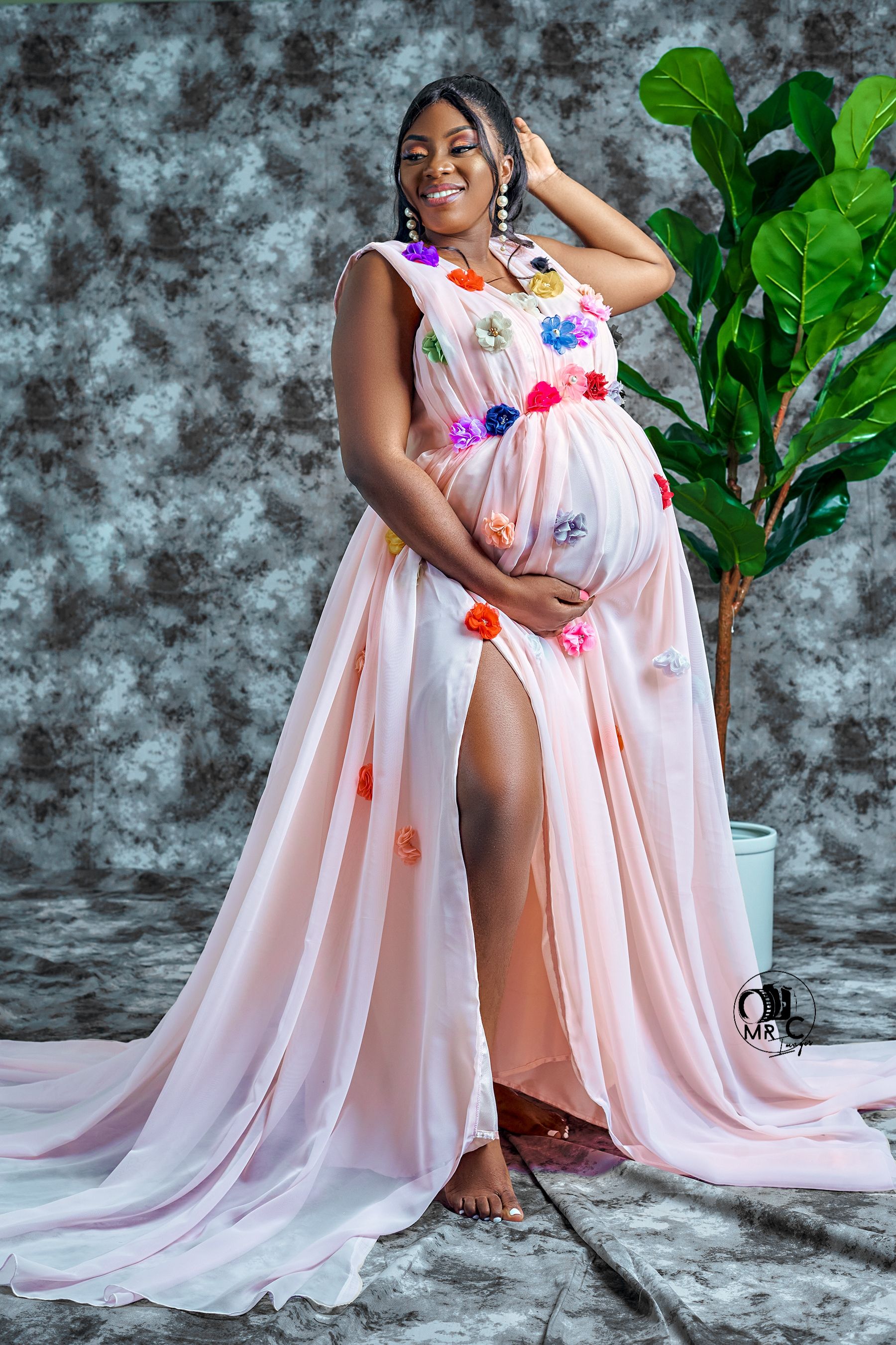 Maternity photo with a flowing pink floral dress and green plant decor against textured grey backdrop.