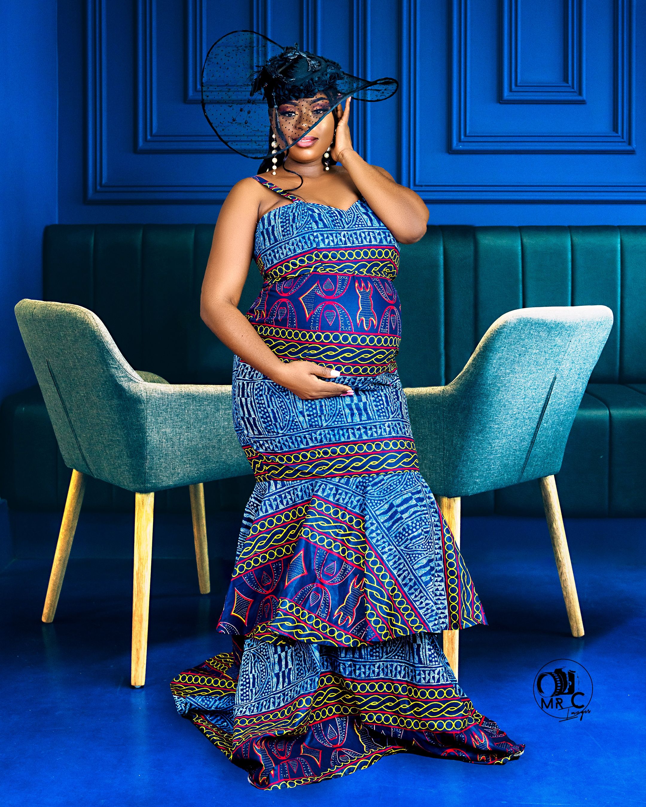 Maternity photoshoot featuring a blue African print gown and teal accent chairs against a royal blue paneled wall.