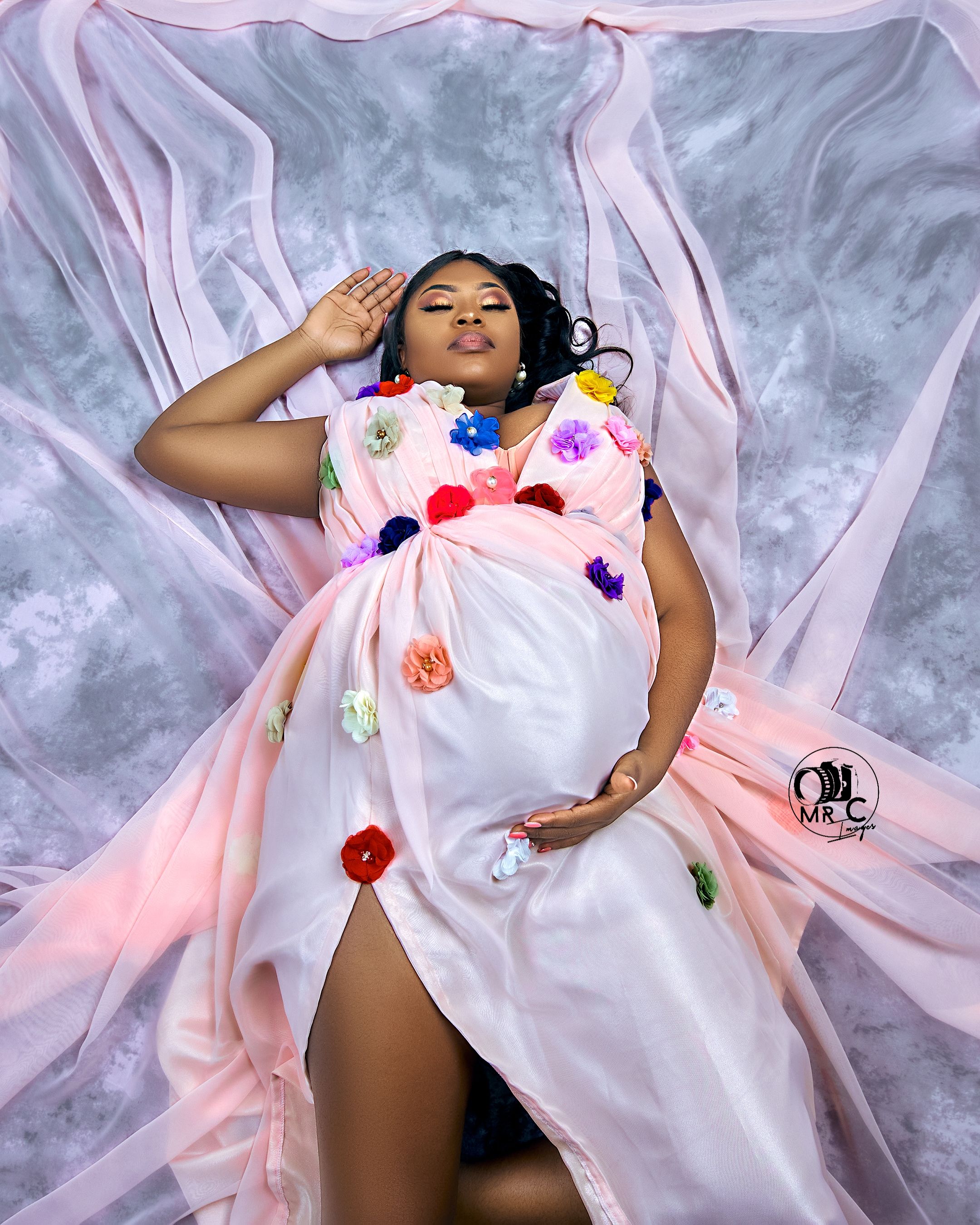 Dreamy maternity photo in pink silk dress with floral accents against marbled background.