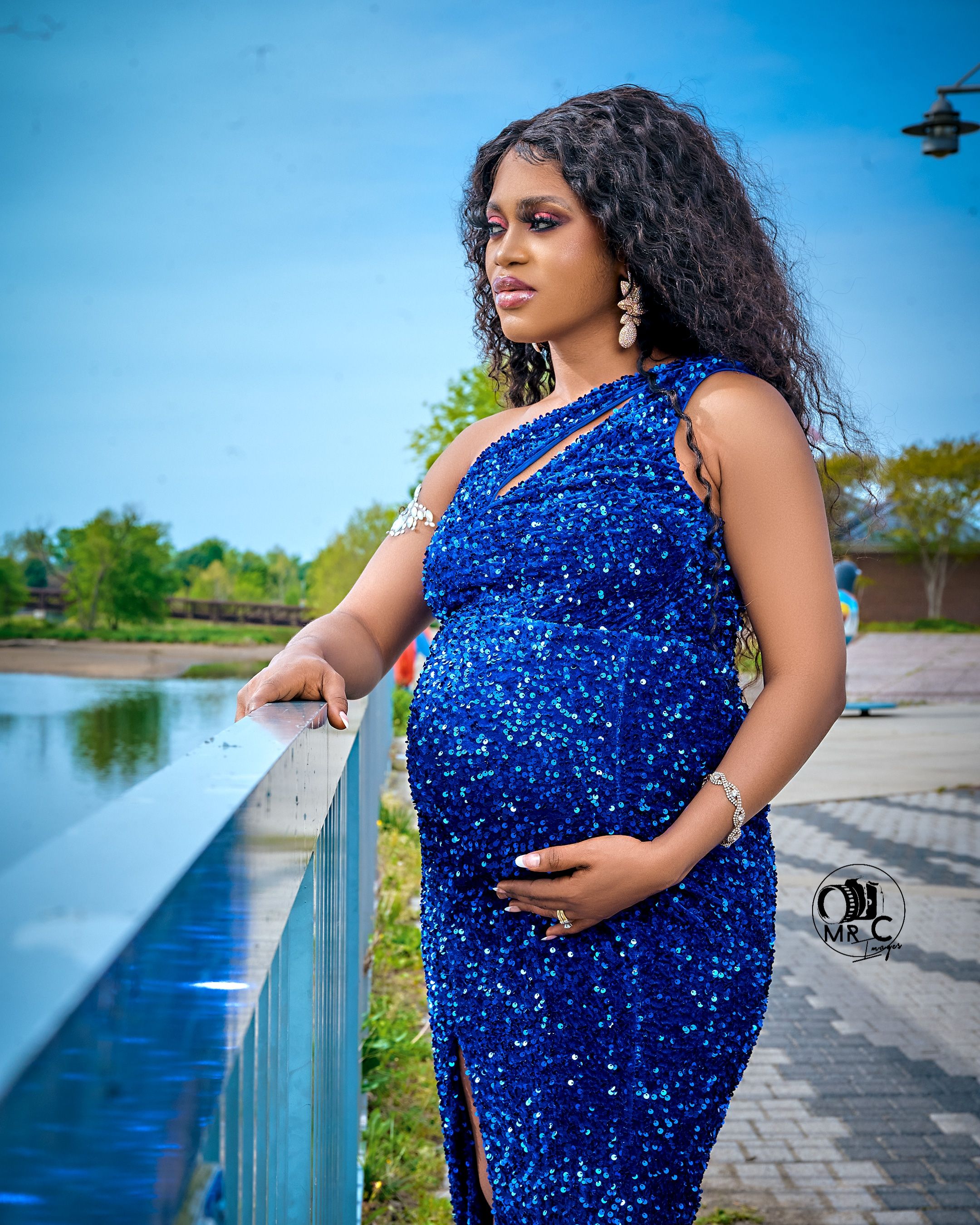Elegant one-shoulder blue sparkly maternity dress photographed by waterfront with scenic lake view.