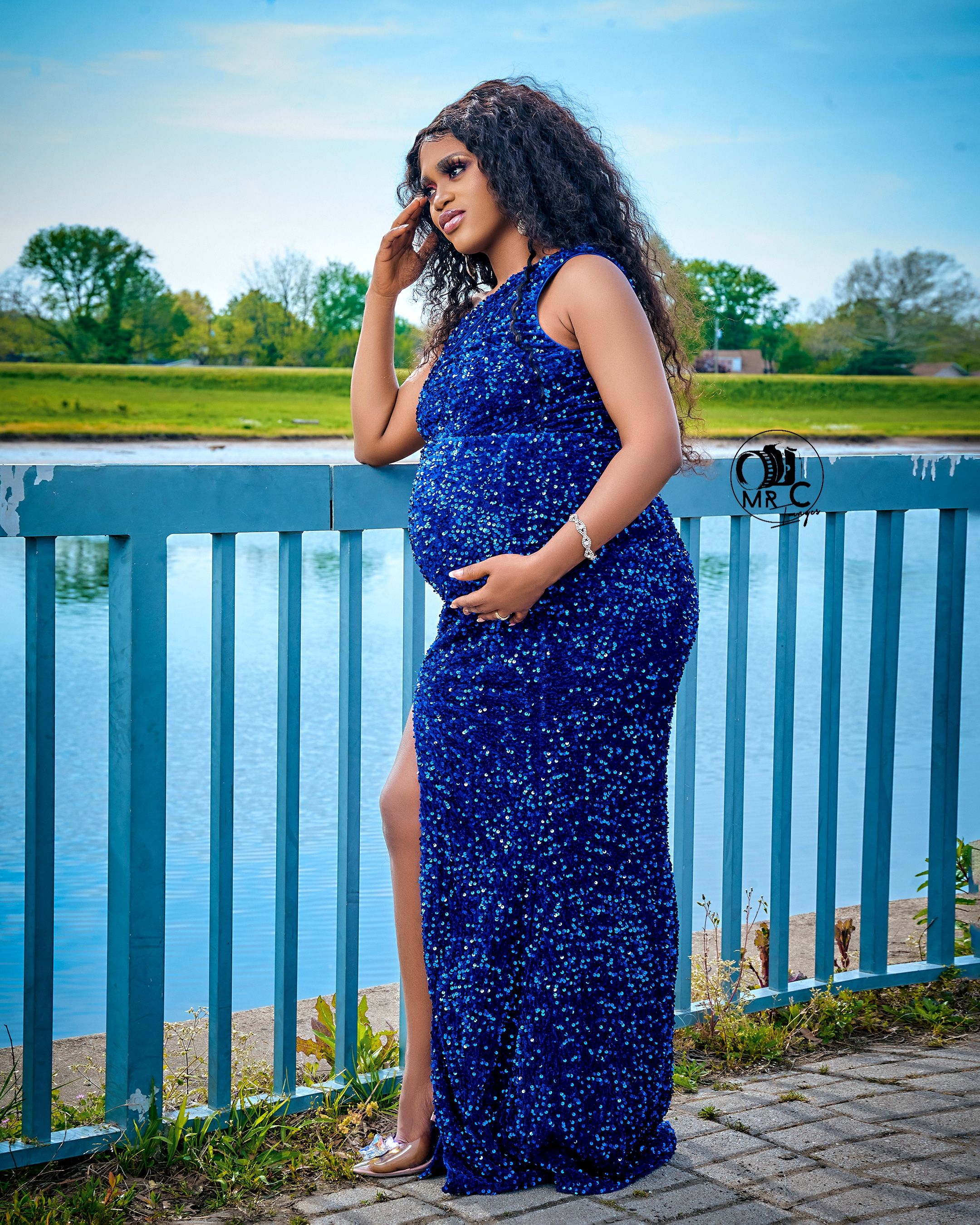 Stunning royal blue sequined maternity gown with side slit photographed against waterfront railing and scenic backdrop.