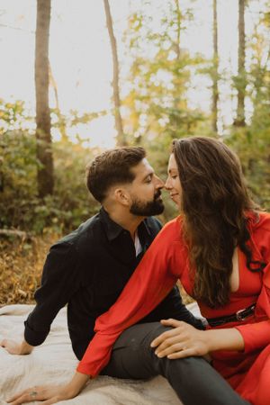 Romantic couple in black and red attire sharing intimate moment on blanket in sunlit forest.