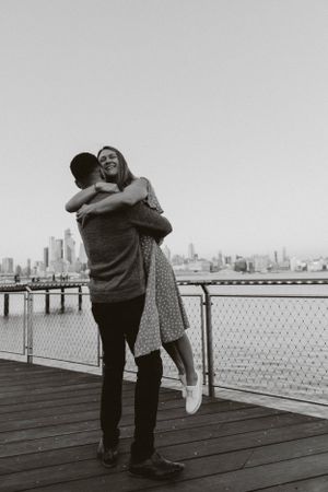A couple sharing a playful moment on a pier with a city skyline in the background.