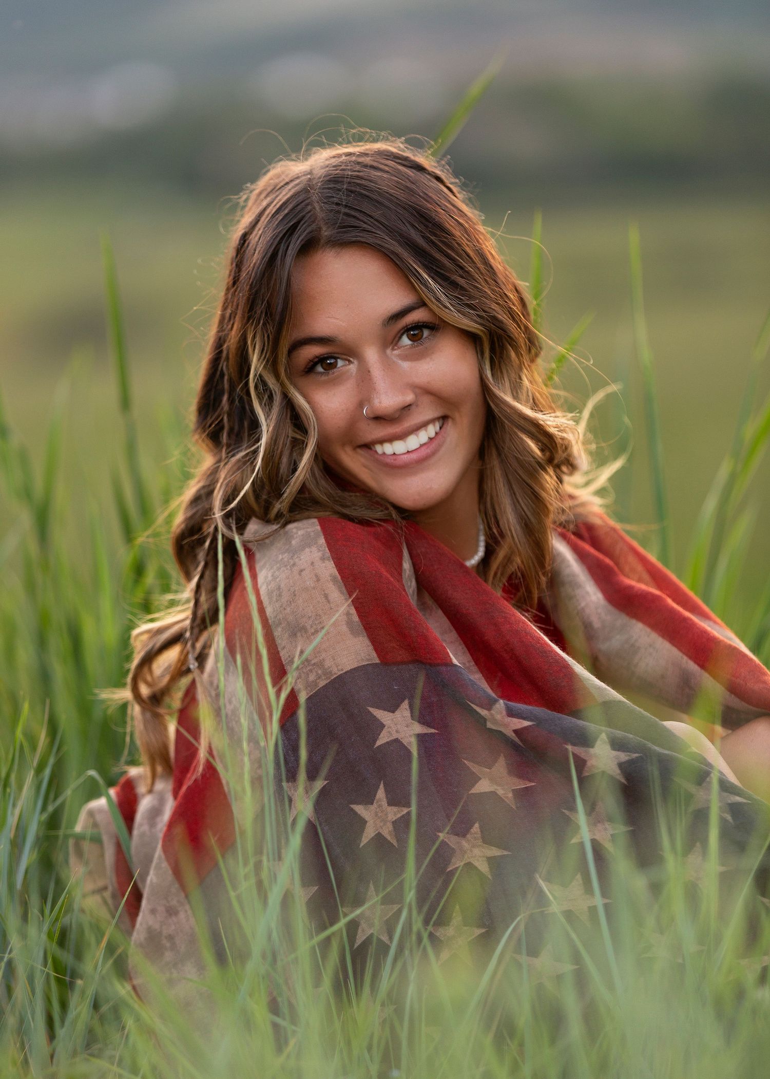 Senior girl sitting in tall grass wrapped in American flag scarf smiling at camera