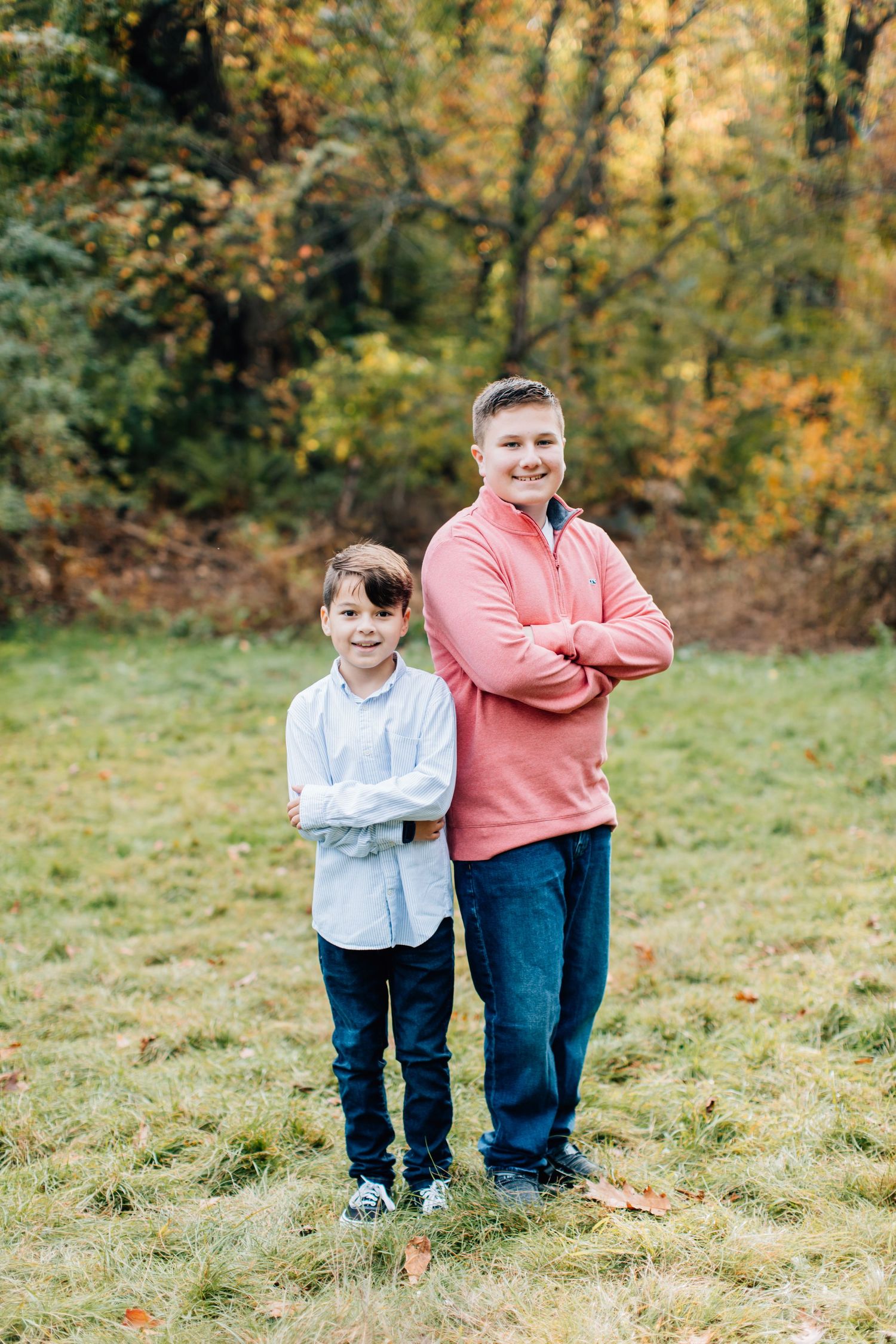 Two people pose casually in a green field surrounded by autumn trees.