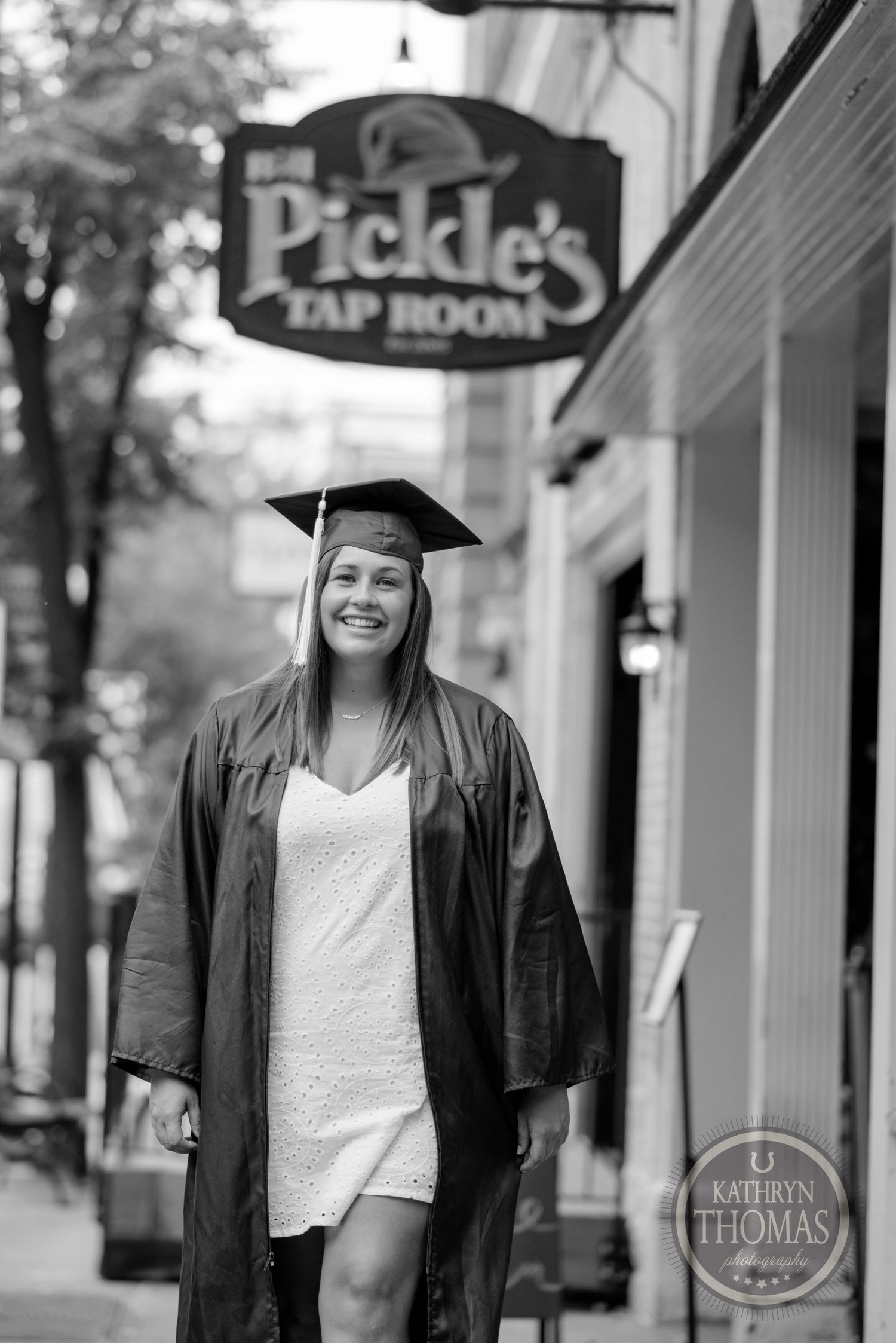 Black and white photo of graduate in cap and gown standing by Pickle's storefront sign.