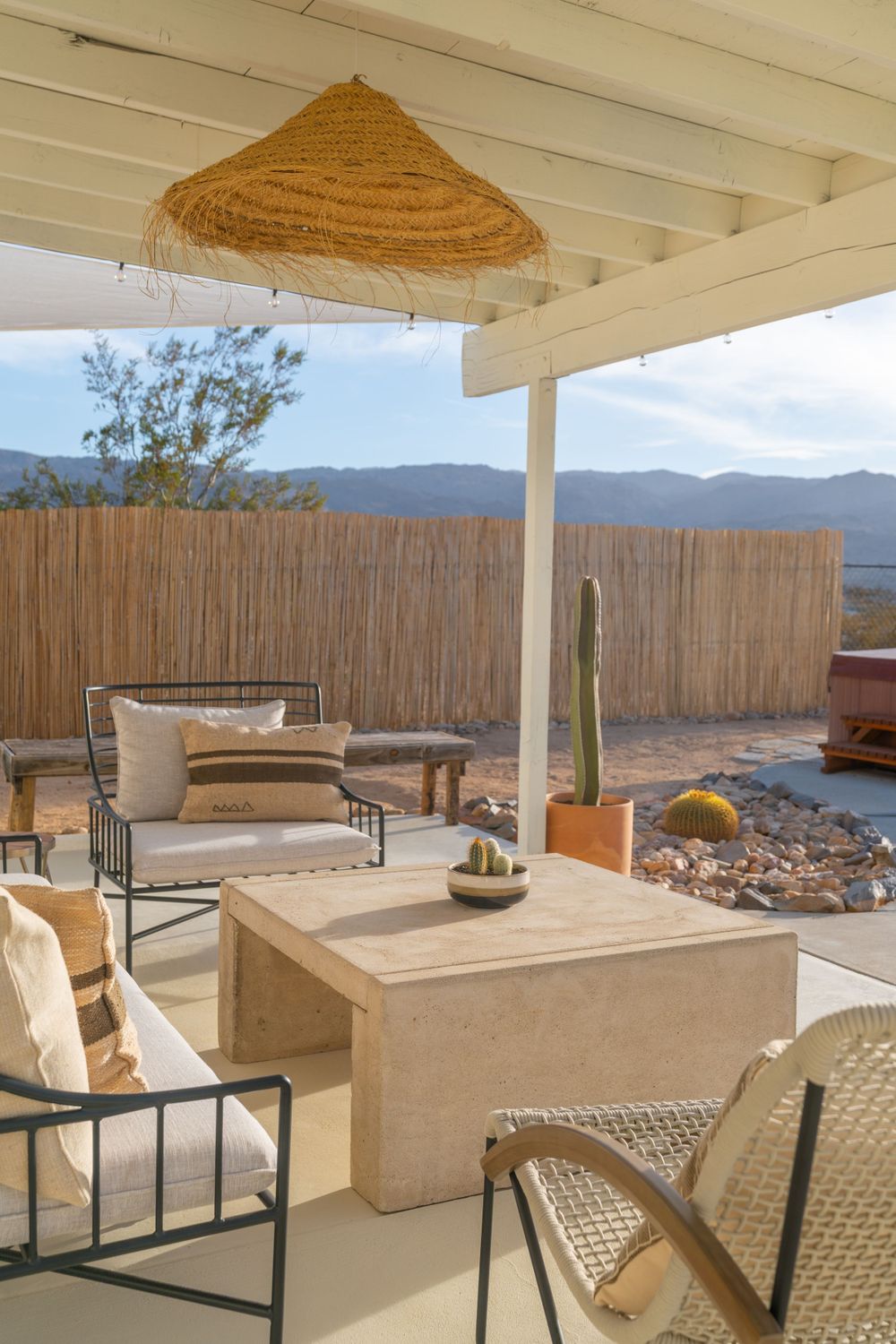 Desert patio with woven umbrella, modern furniture, and mountain views through bamboo privacy screen.