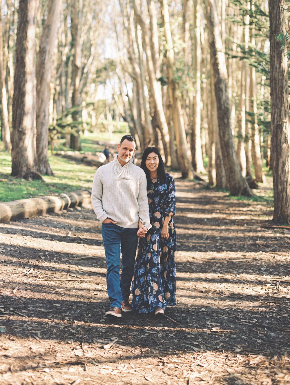 A couple in coordinated outfits stands together on a tree-lined path during a photo session.
