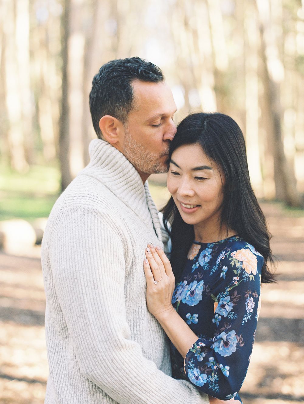 A tender moment between a couple in a white sweater and floral dress against a forest backdrop.