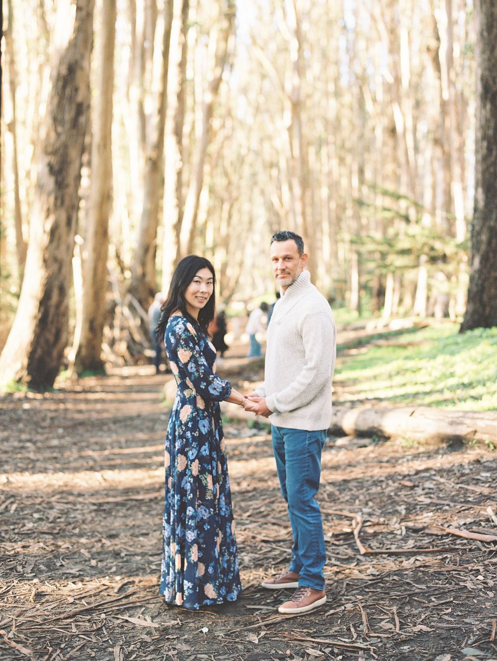 Couple in coordinated outfits poses together in sunlit forest setting.