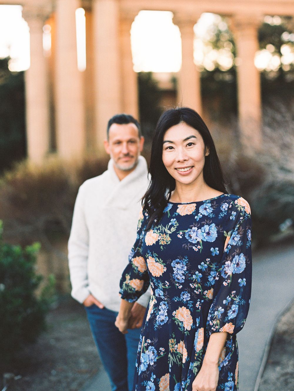 Stylish couple poses outdoors, woman in floral maxi dress with man in casual wear behind her.