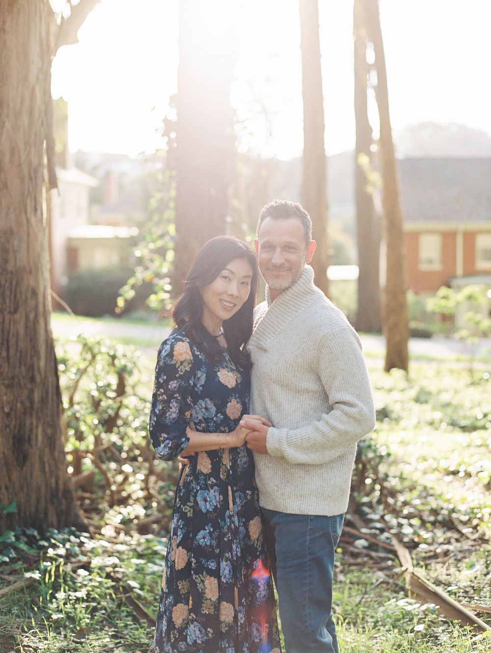 A couple poses together in a sun-dappled forest wearing coordinated casual outfits.