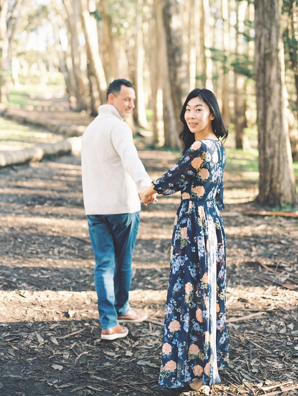 A romantic couple holds hands while walking through a scenic wooded area, turning to look at each other.