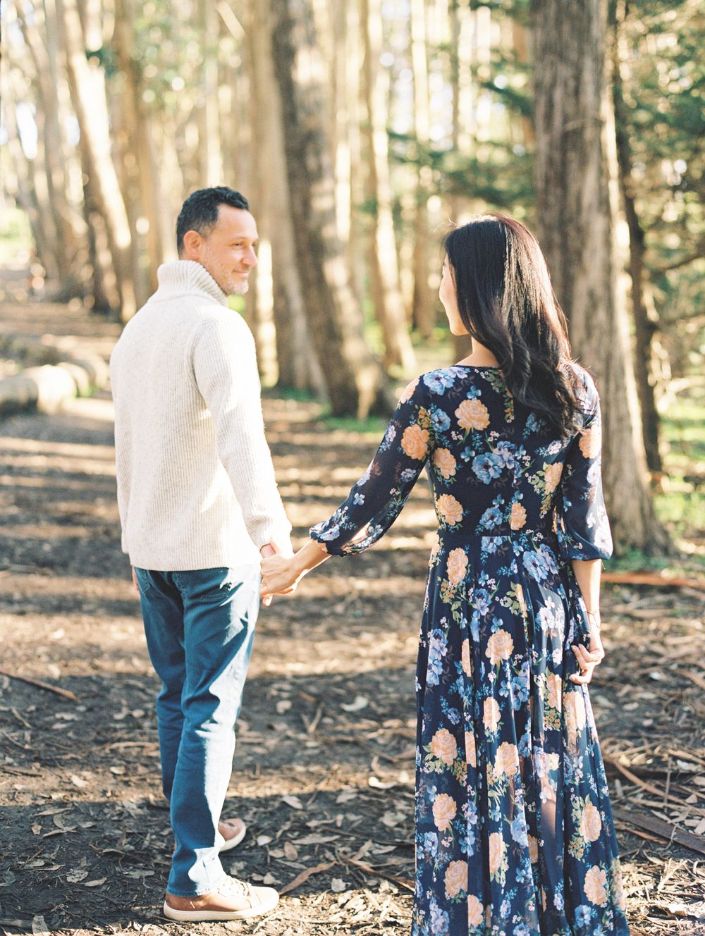 A couple holds hands while walking along a sunlit forest path with tall trees in the background.