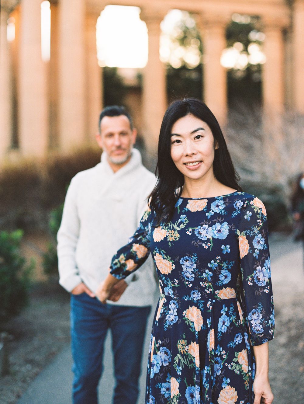 A couple poses in front of stone pillars with woman in blue floral dress and man in cream sweater.