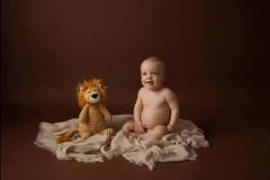 Simple studio portrait with baby sitting next to plush lion toy on brown backdrop.