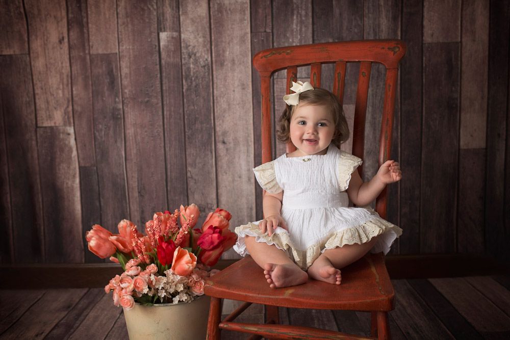Small child in white dress sits on wooden chair next to orange tulips against rustic backdrop.