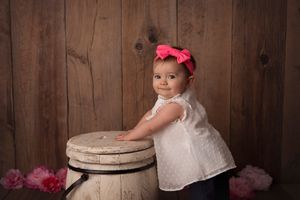 Child in white dress leans against vintage barrel with pink bow accessory.