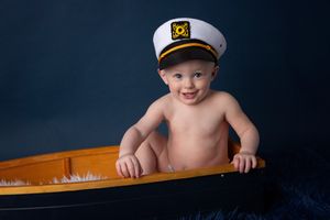 Child wearing captain's hat poses playfully in toy boat against dark blue background.