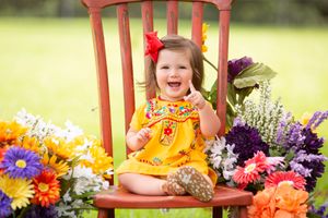 Child in yellow dress sits on pink chair surrounded by colorful summer flowers.
