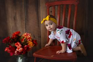 Playful pose on a red wooden chair next to orange flowers in vintage setting.